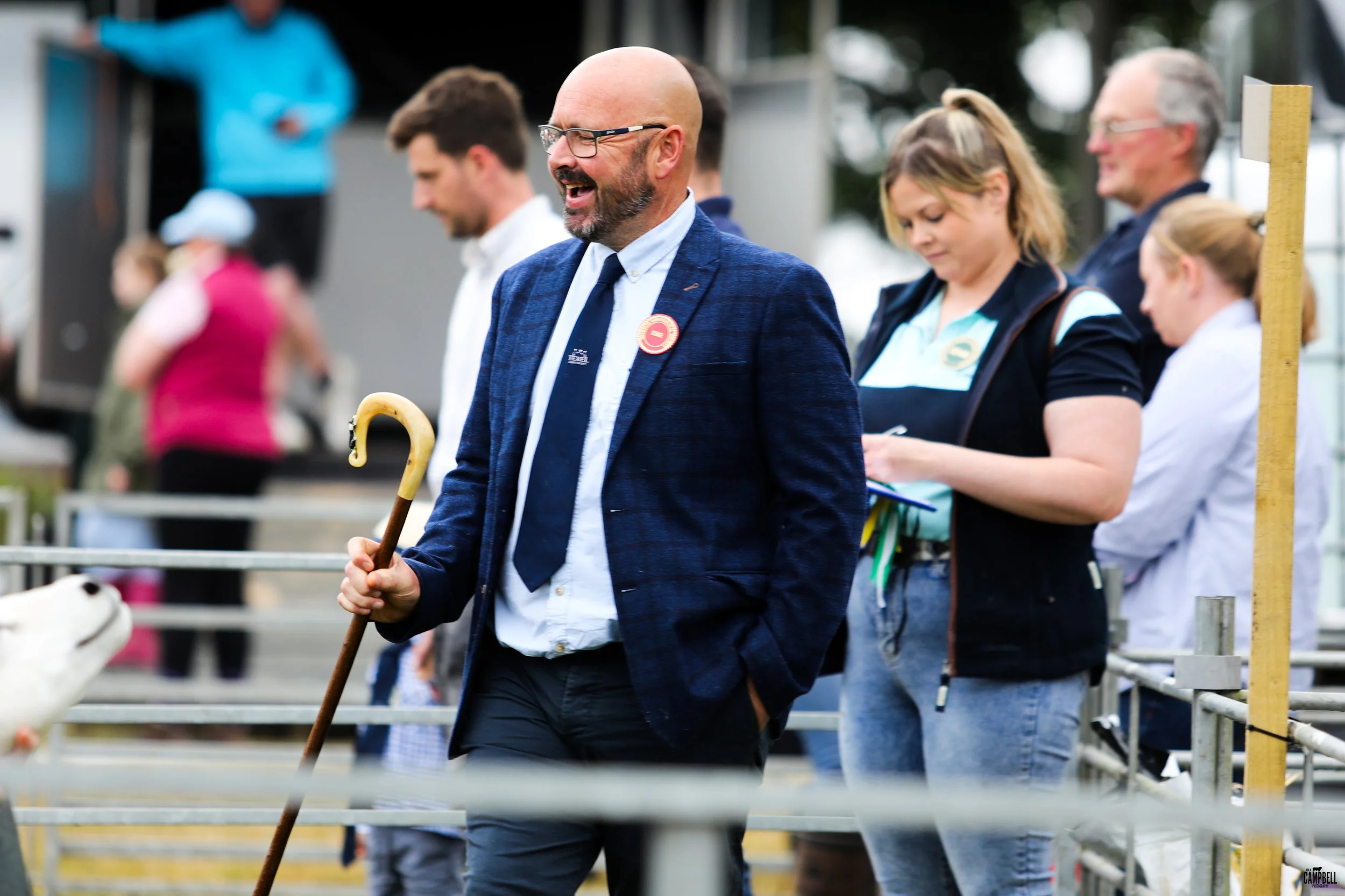 A man in a blue blazer and glasses walking with a cane at an outdoor event, with several people standing nearby, some taking notes or looking down.