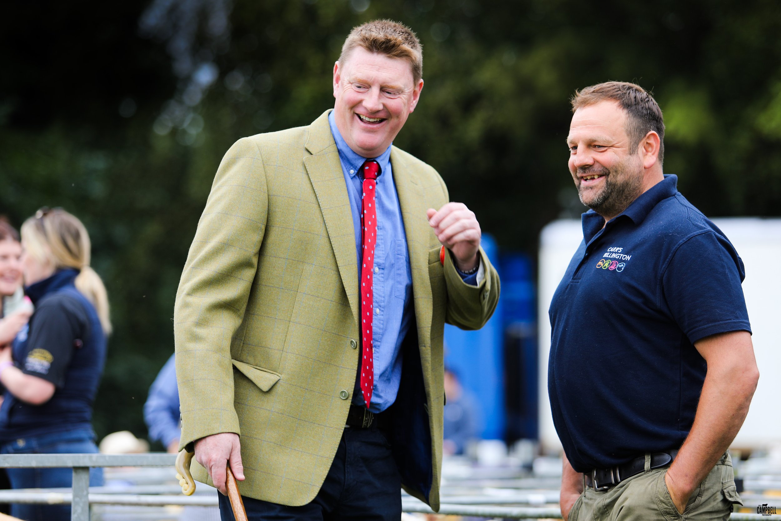 Two men smiling and talking outdoors, one wearing a yellow blazer and the other a blue shirt with logo, in a grassy area with people in the background.