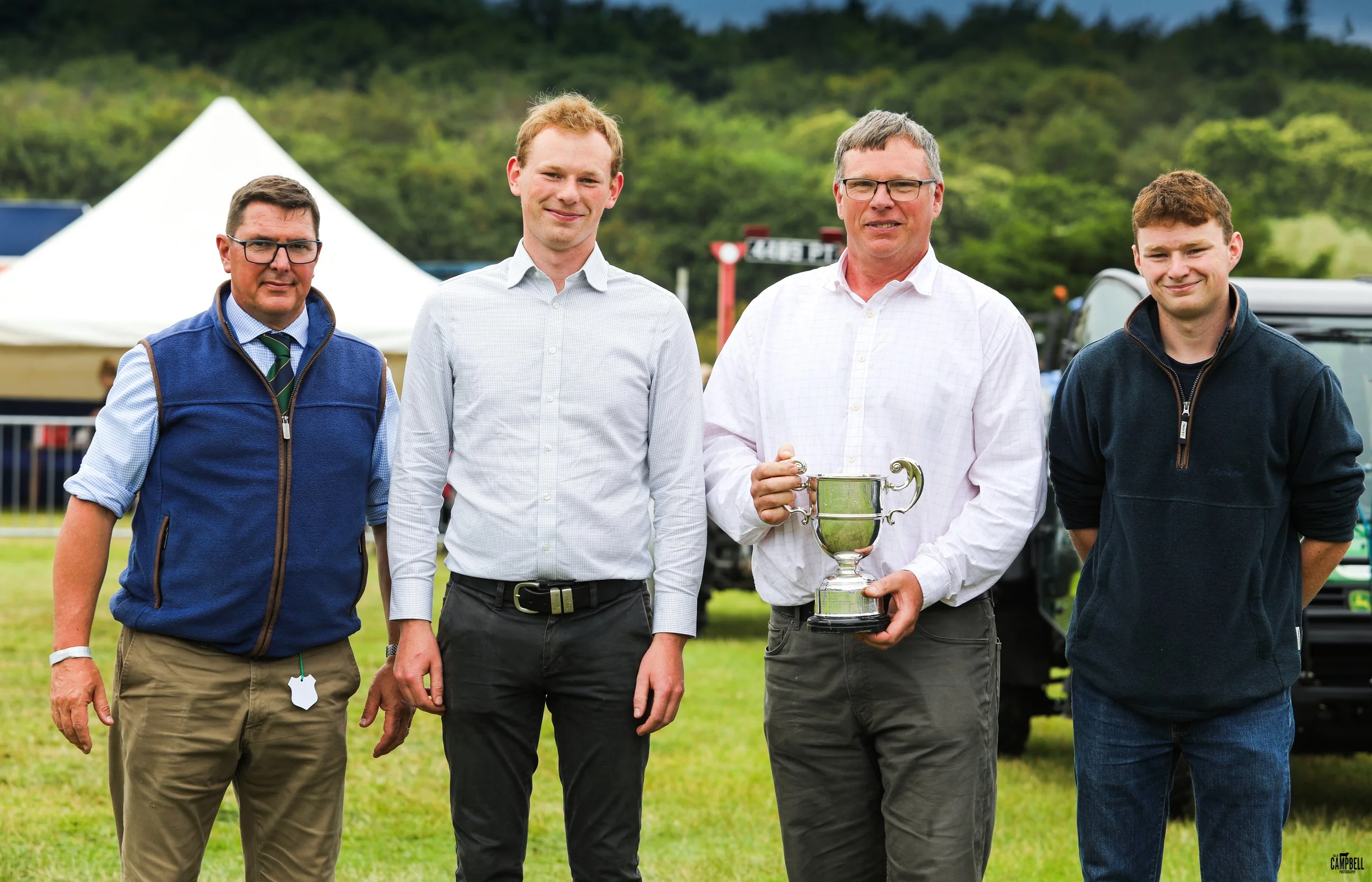 Four men standing outdoors on grass, with tents and trees in the background. The second man from the right is holding a trophy. All are smiling.