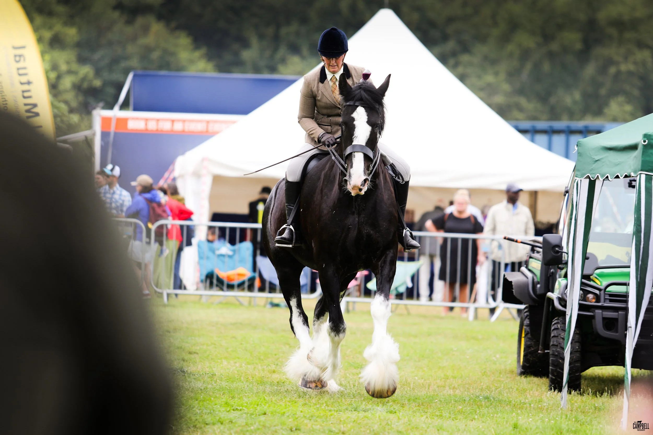 A person riding a black and white horse at an outdoor event, with tents and people in the background.