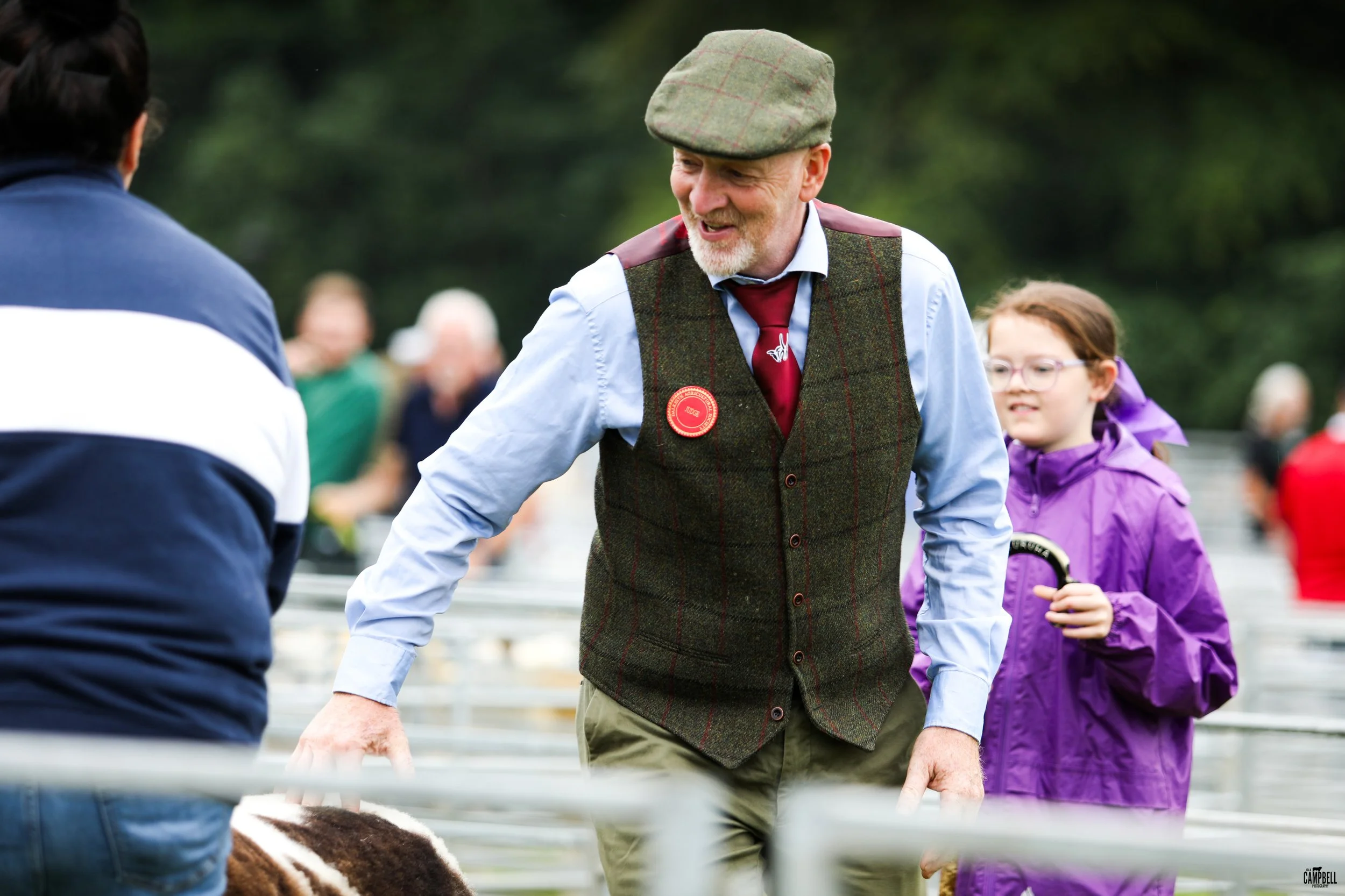 An older man with a white beard wearing a flat cap, a tartan vest with a 'Judge' badge, and a light blue shirt, is smiling and talking to a person whose back is to the camera. The background has people and trees, suggesting an outdoor event.