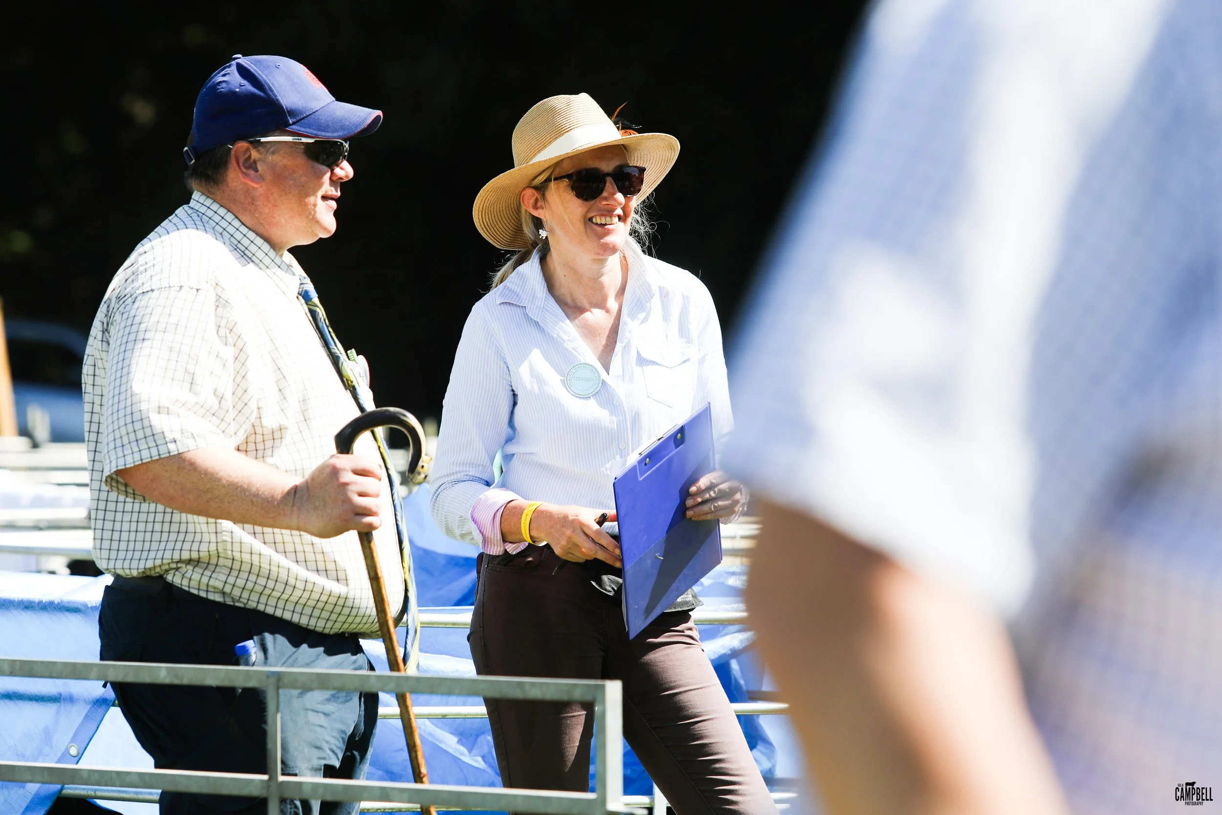 Two people conversing outdoors, one man holding a cane and wearing a hat and sunglasses, and one woman holding a clipboard, wearing a hat and sunglasses, both dressed in casual clothing, with tons of sun.