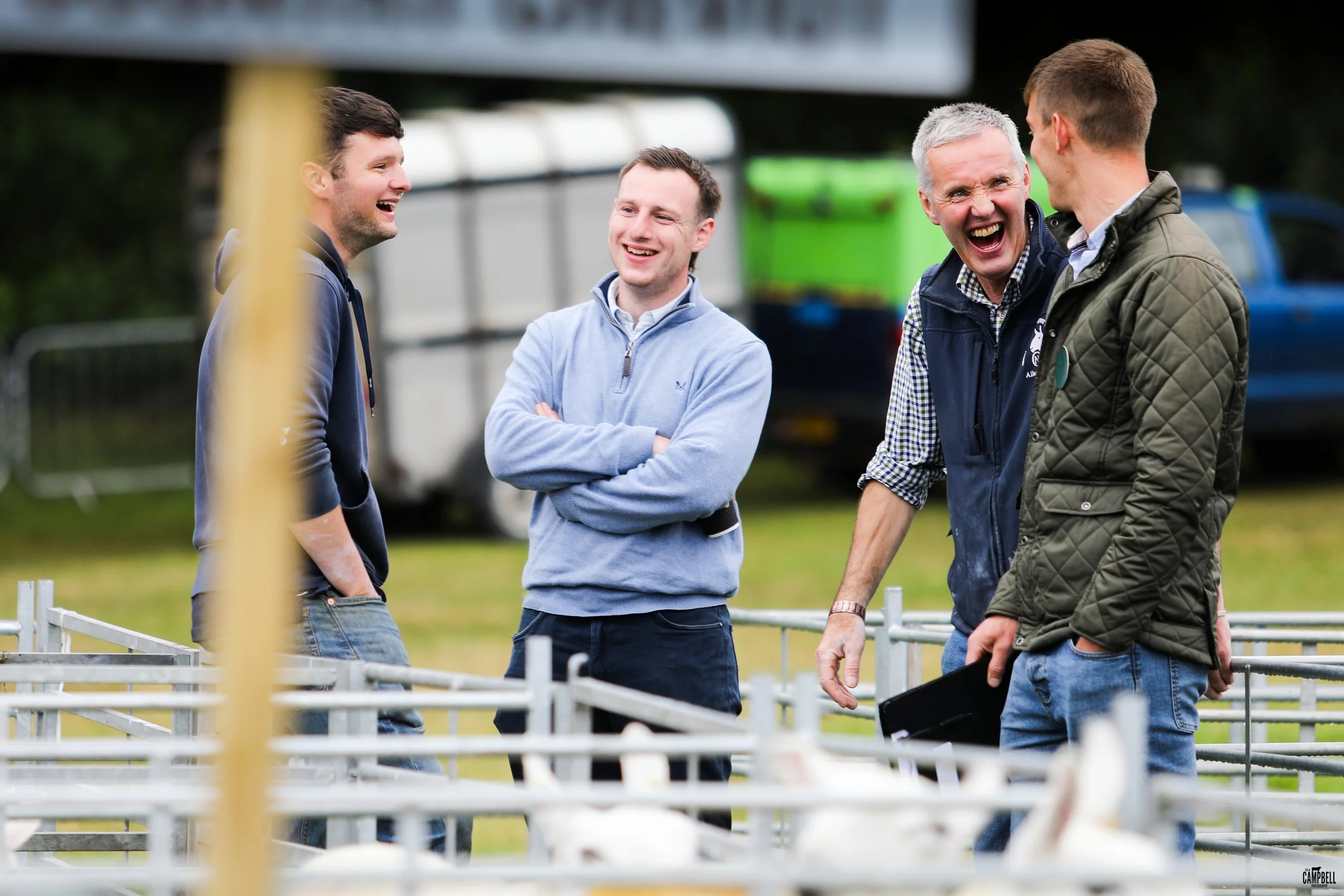 Four men standing outdoors, laughing and talking, near a white fence, with a background of parked cars and greenery.