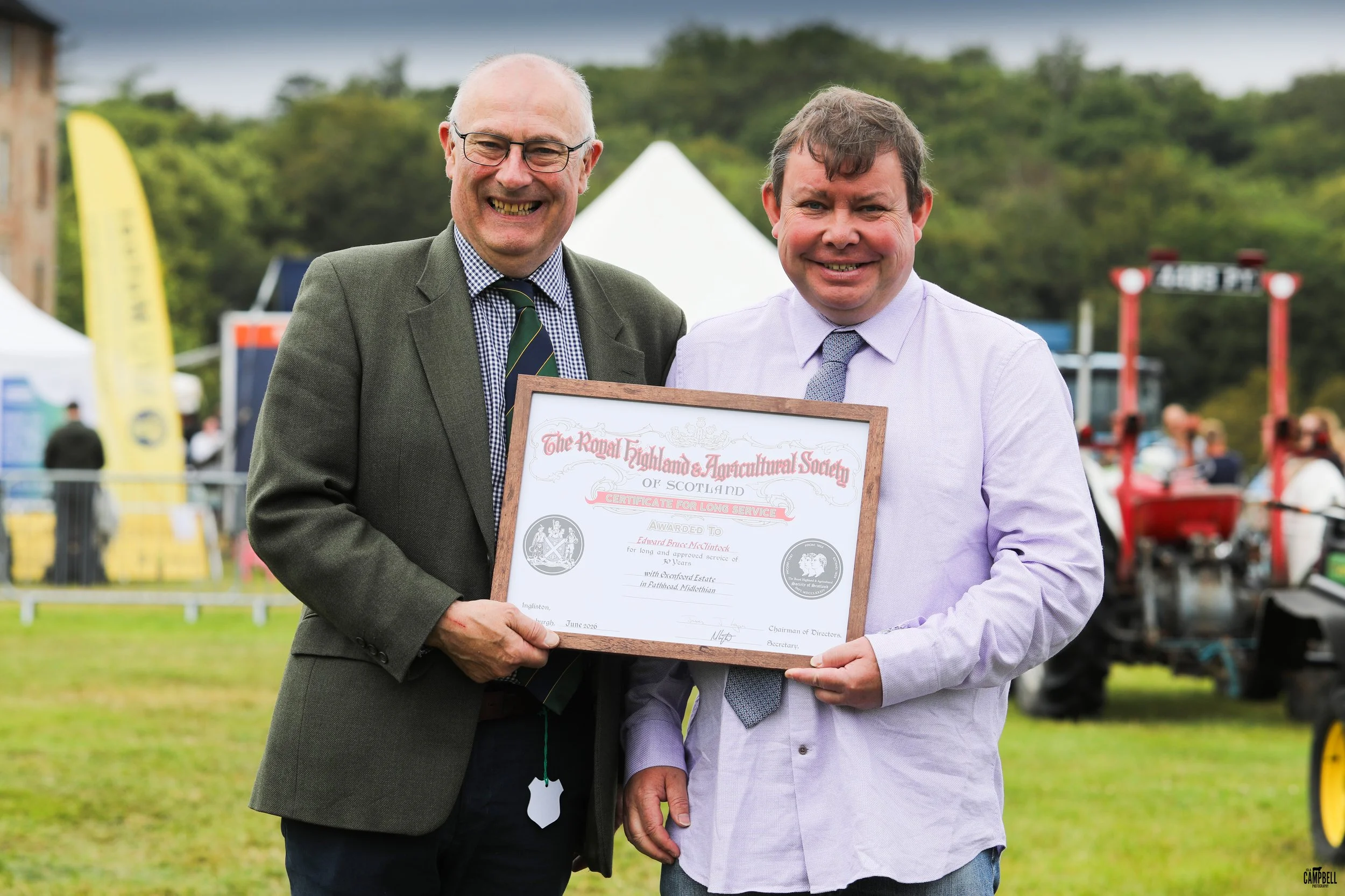 Two men smiling at an outdoor event, one holding a framed certificate from the Royal Highland and Agricultural Society of Scotland, with tents and people in the background.