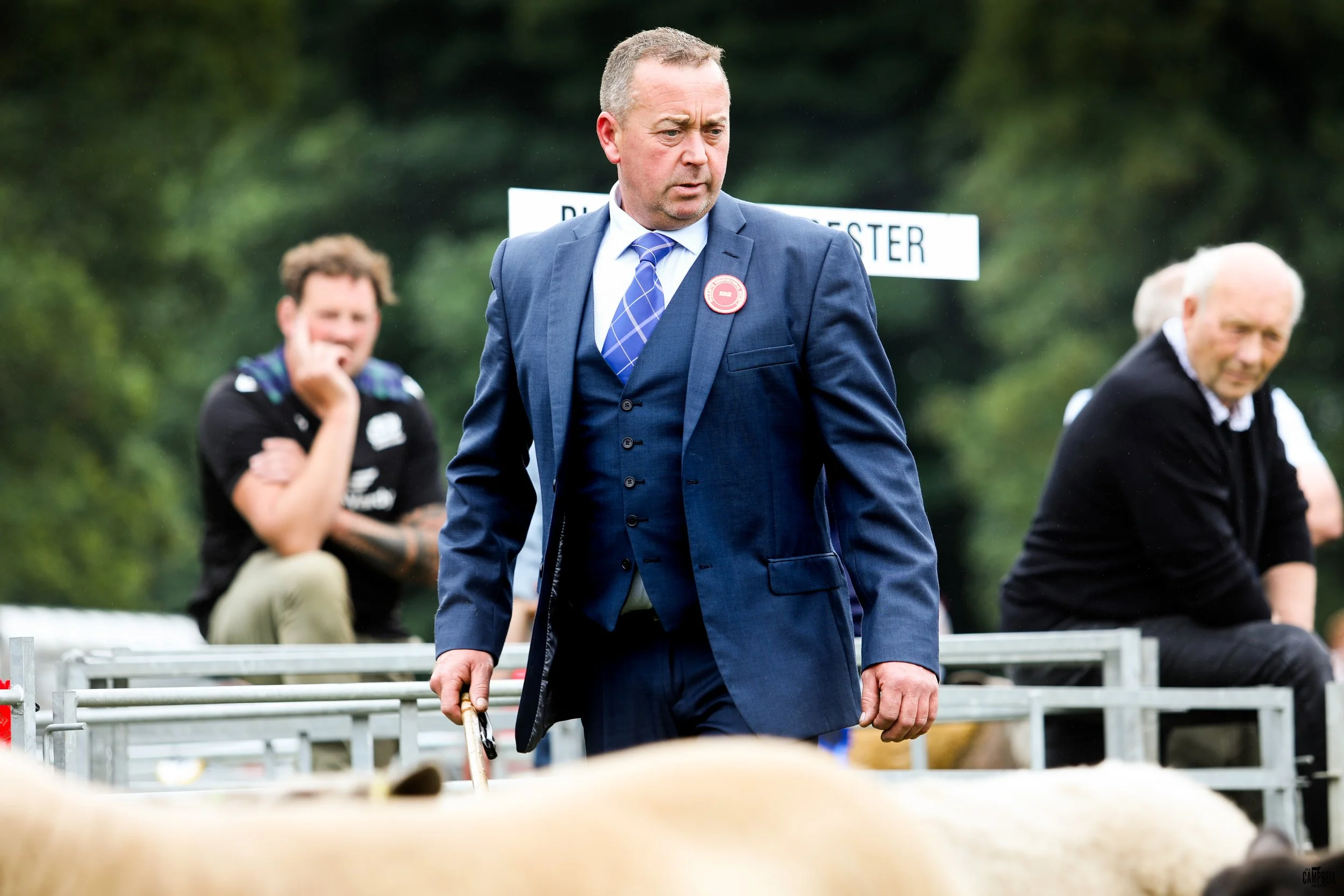 A man in a blue suit and tie walking with a cane at an outdoor event, with two other men in the background, one with a tattoo on his arm and the other with white hair, all near a white fence and trees.