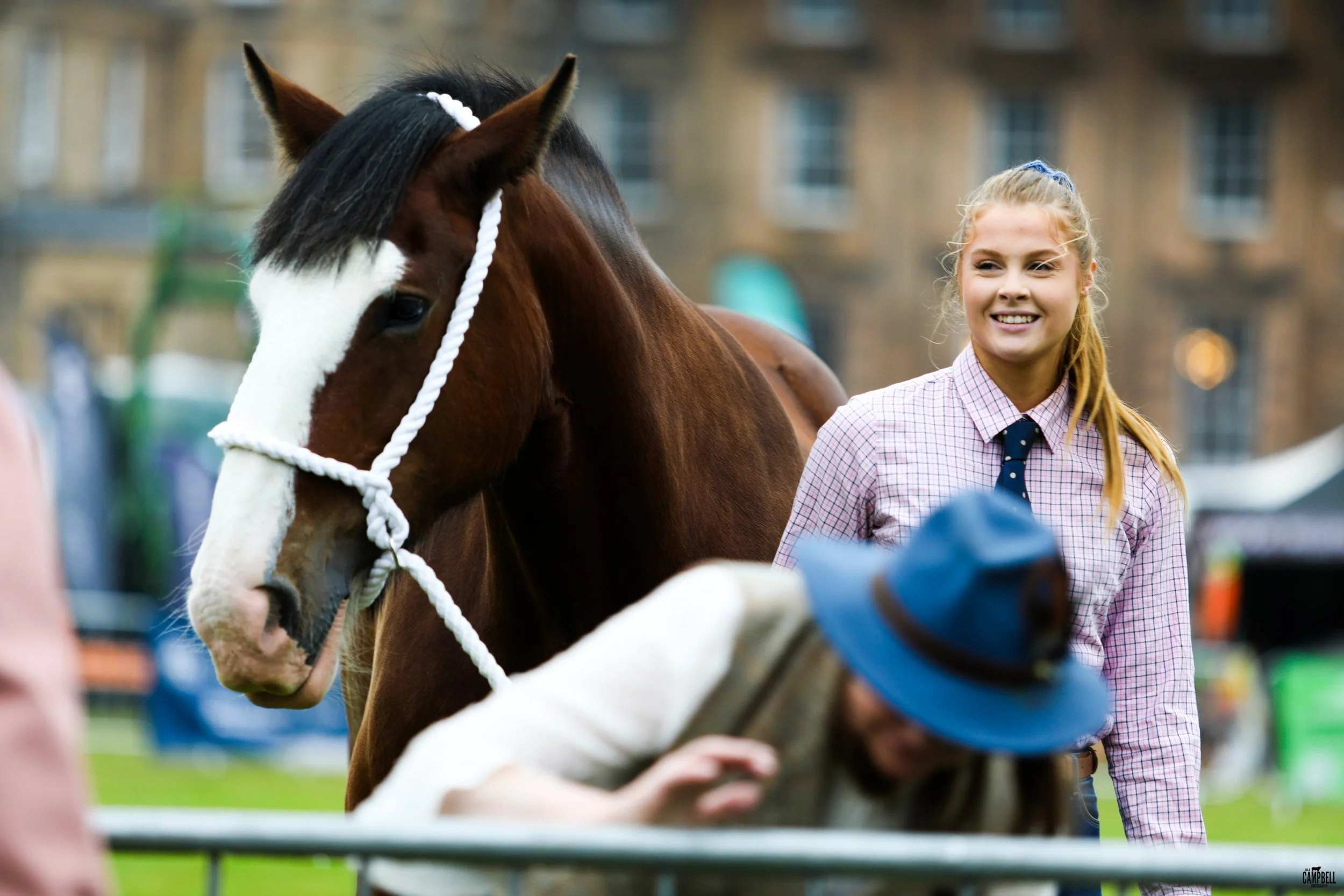 A young girl with blonde hair tied in a ponytail smiling and standing next to a brown and white horse with a white rope halter, in an outdoor setting with buildings in the background.