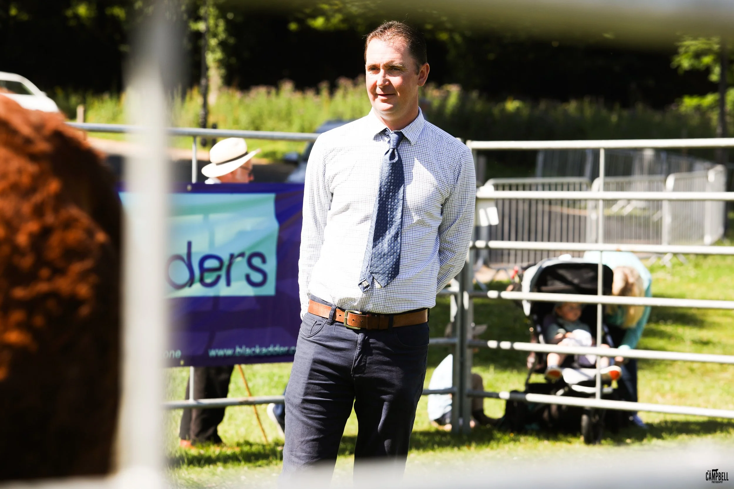 A man in a checkered shirt and tie standing outdoors near a white fence, with a grassy area, a banner, and children in the background.