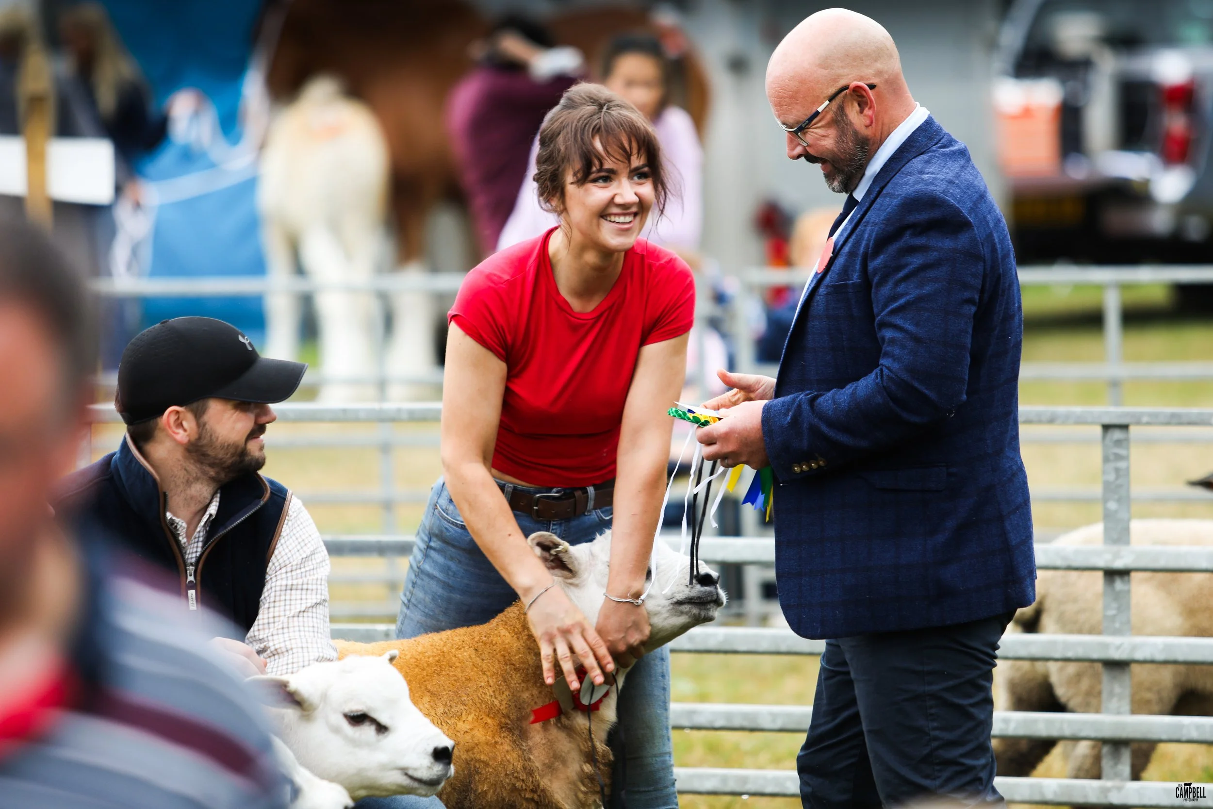 A young woman in a red shirt smiling at a man in a blue blazer at a rural fairground, with a sheep, goats, and other people in the background.