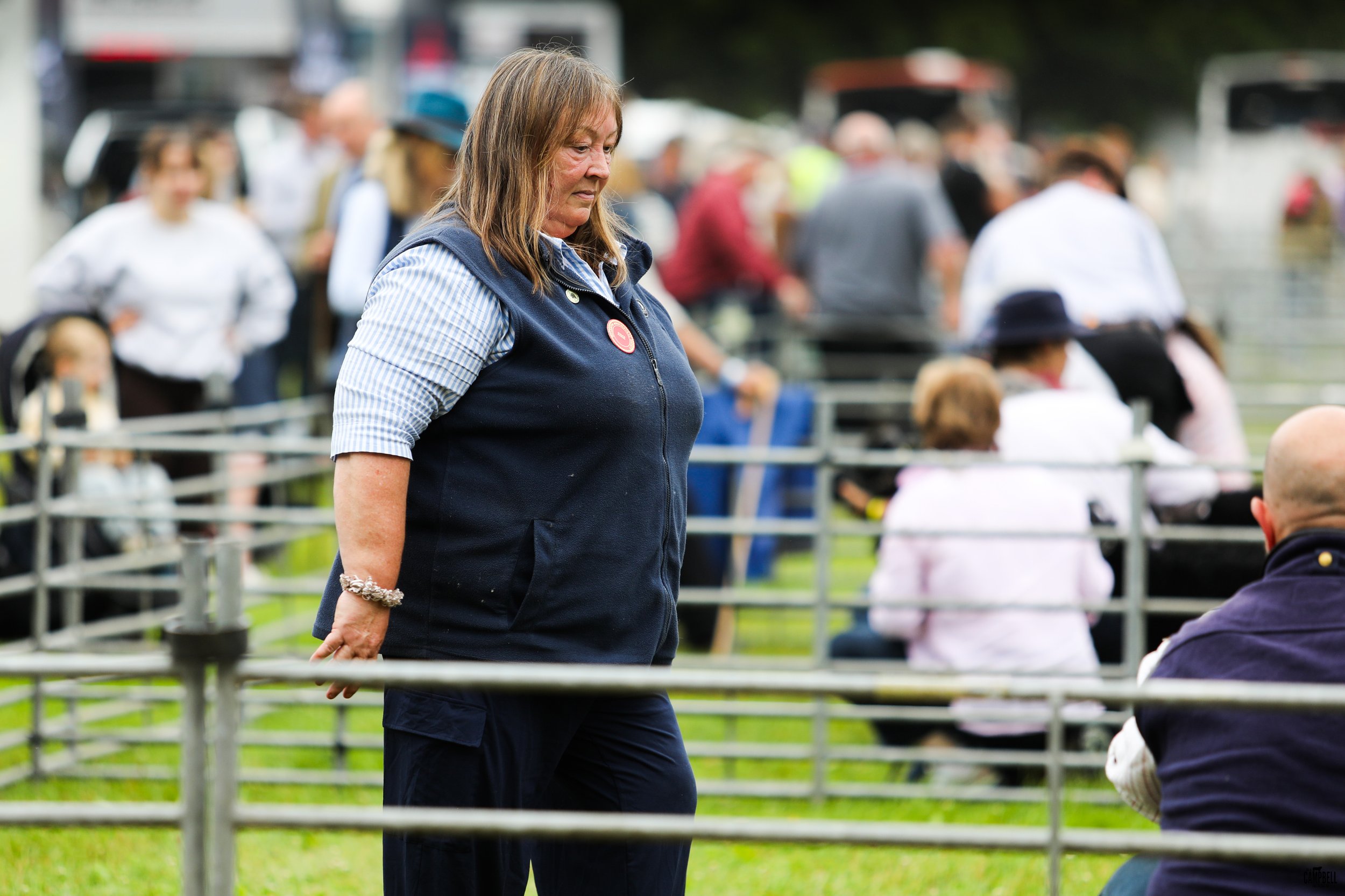 A woman with shoulder-length brown hair wearing a dark vest and a striped shirt standing outdoors in front of a crowd of people and metal fencing.