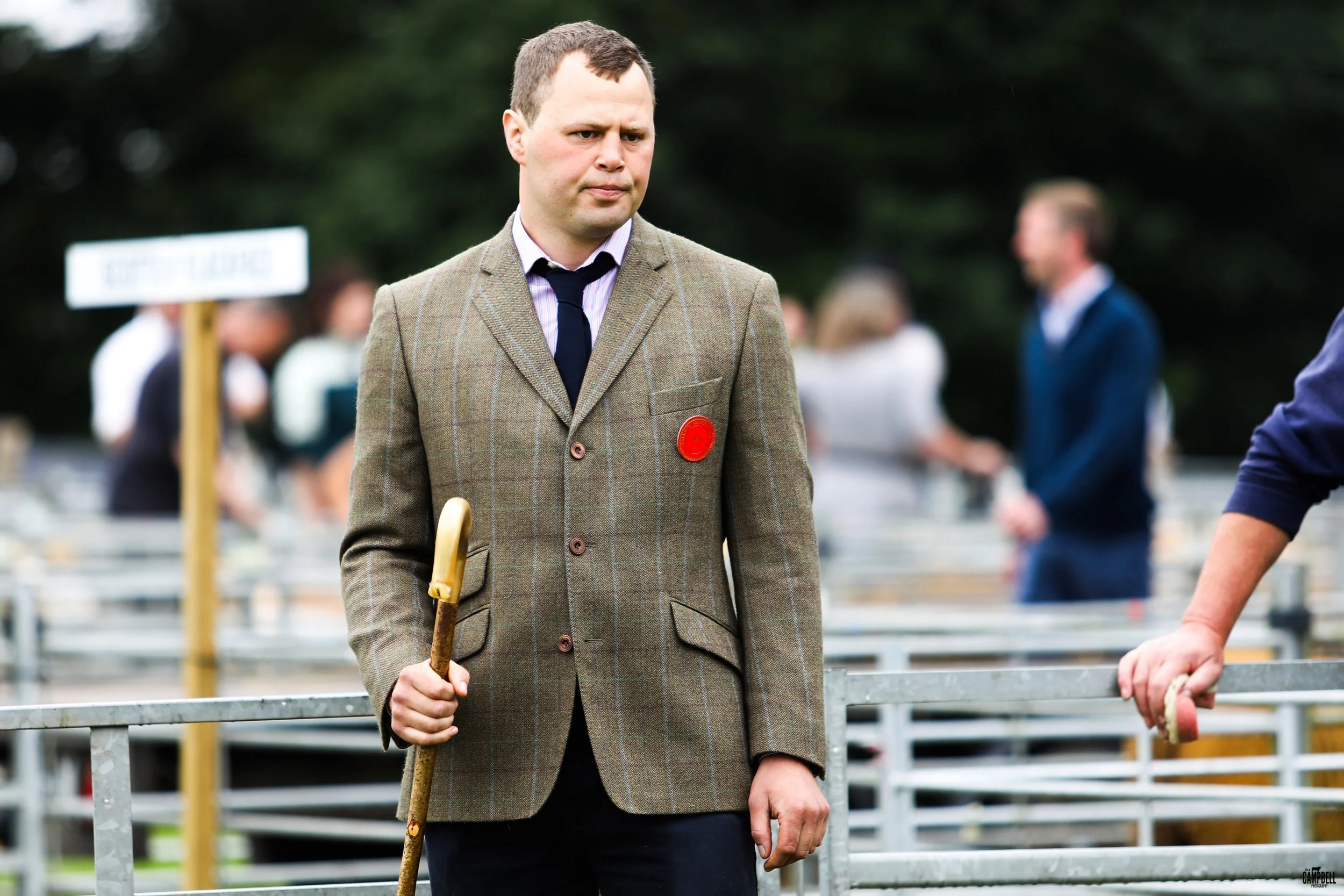 A man in a brown checkered blazer holding a stick, standing outdoors at an event with blurred people in the background.