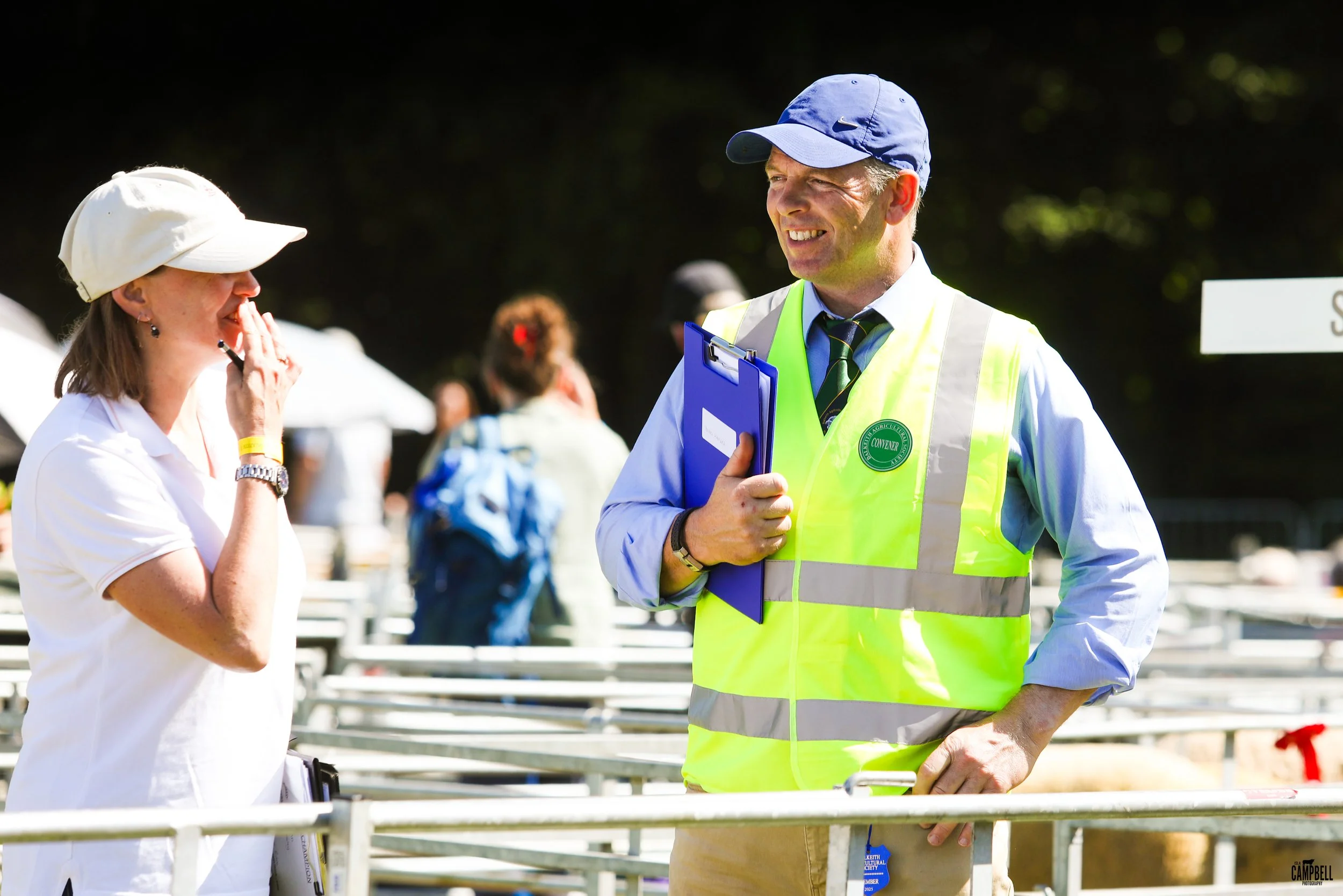 A man wearing a blue cap and a high-visibility vest talking to a woman with a white hat at an outdoor event.