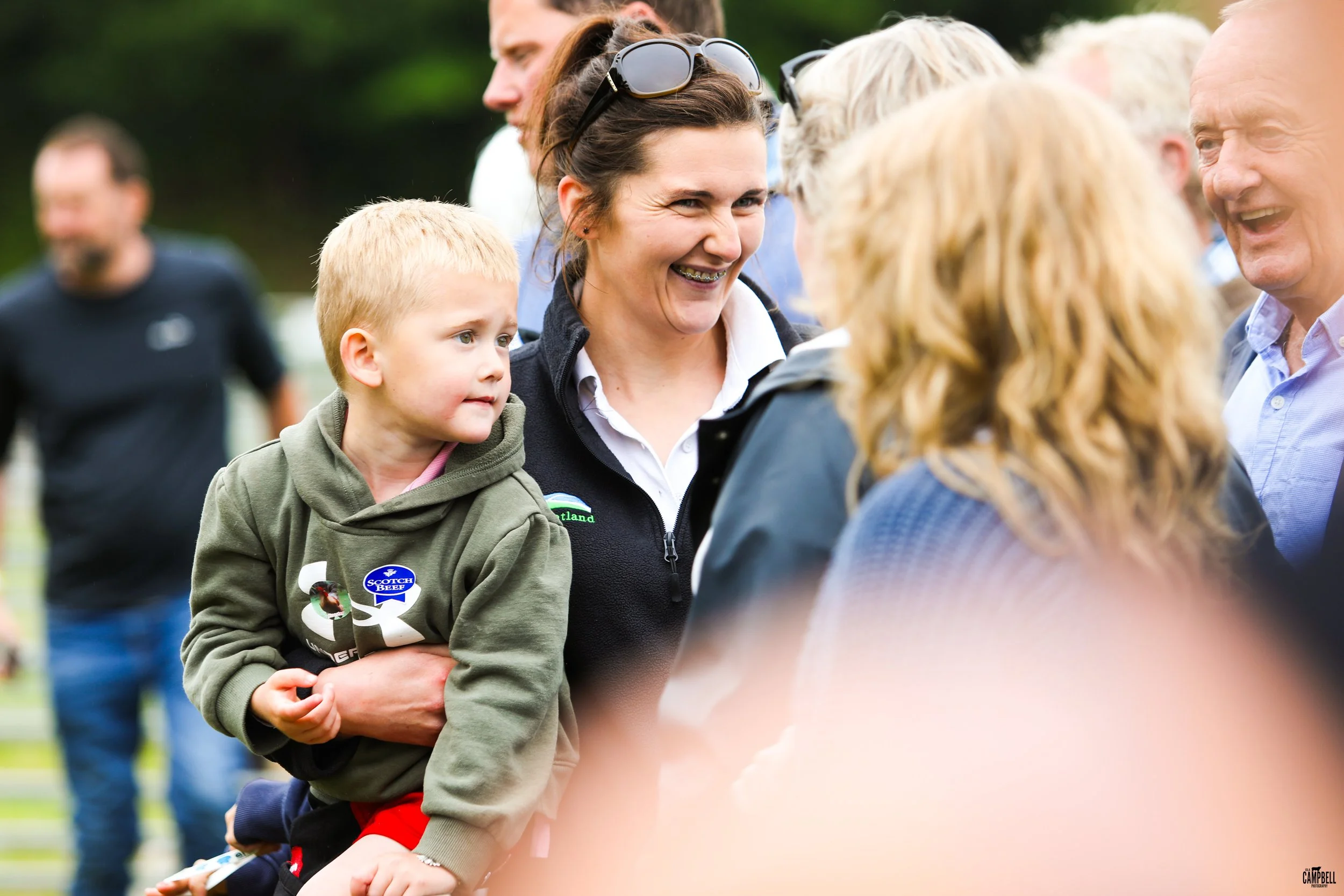 A group of people, including a woman with sunglasses on her head and braces, holding a young boy with blonde hair and a green hoodie, smiling and engaging in conversation at an outdoor event.
