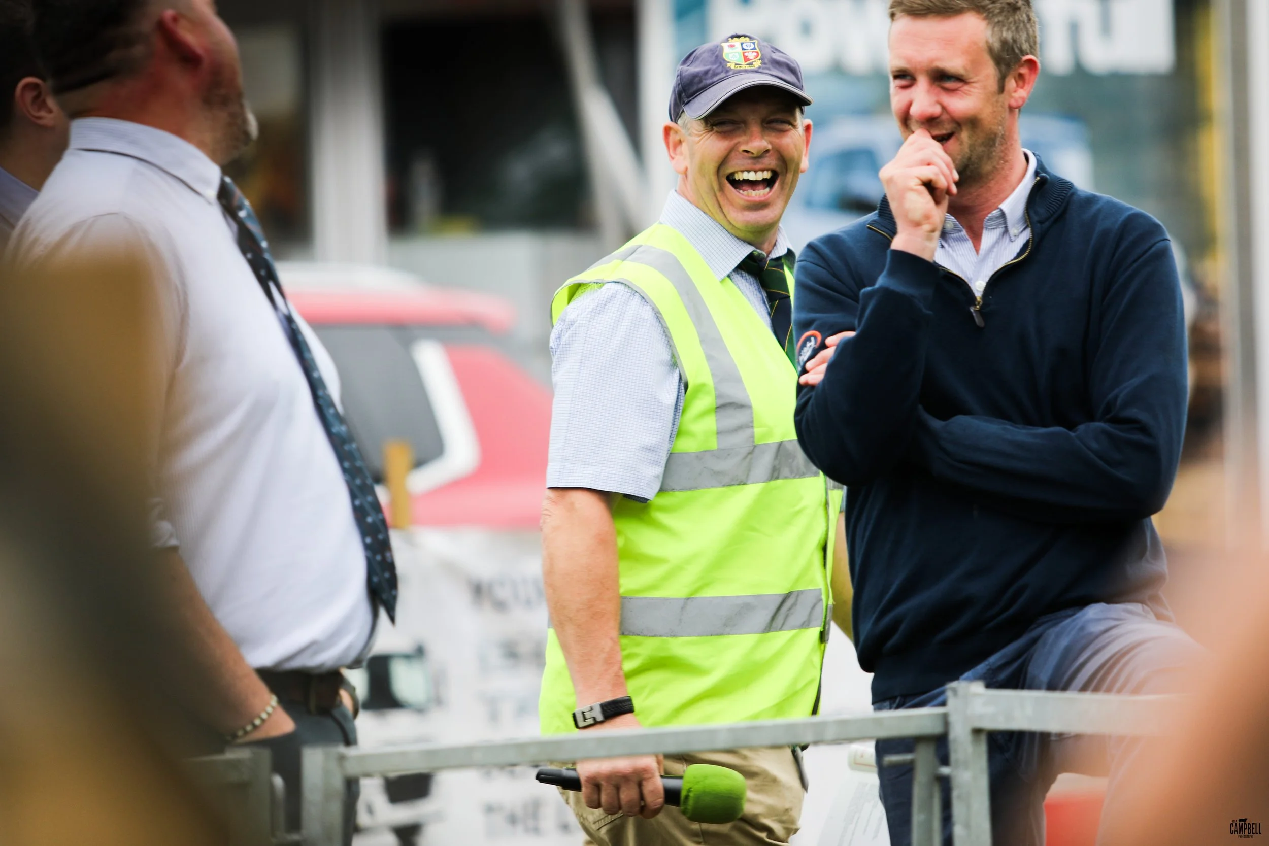 A group of men engaged in conversation outdoors, one man wearing a bright yellow safety vest and a baseball cap, holding a tool, among others dressed in business casual clothing, smiling and laughing.