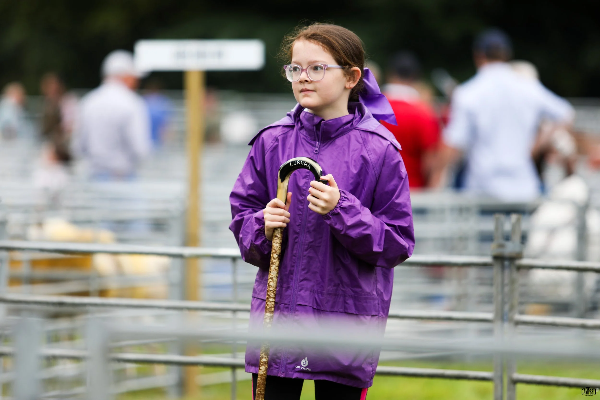 A young girl with glasses wearing a purple rain jacket and holding a stick, standing among metal barriers at an outdoor event on a cloudy day.