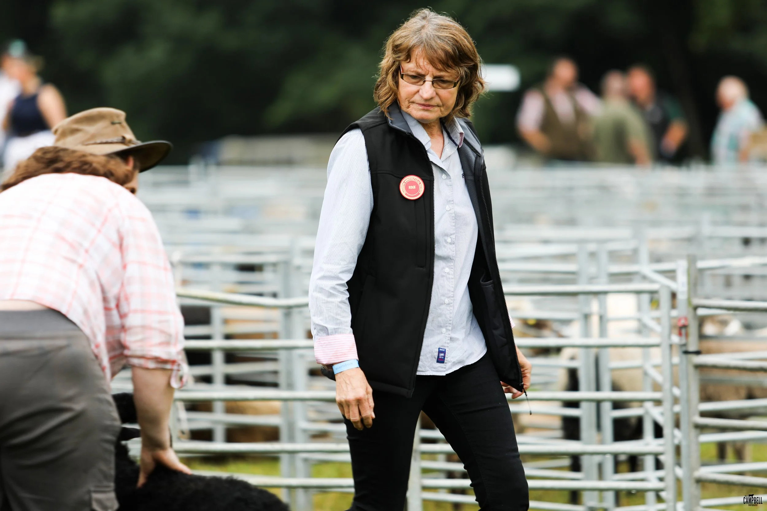 A woman with glasses wearing a black vest over a light blue shirt looking at a dog, with a group of people blurred in the background at an outdoor event with white fencing.