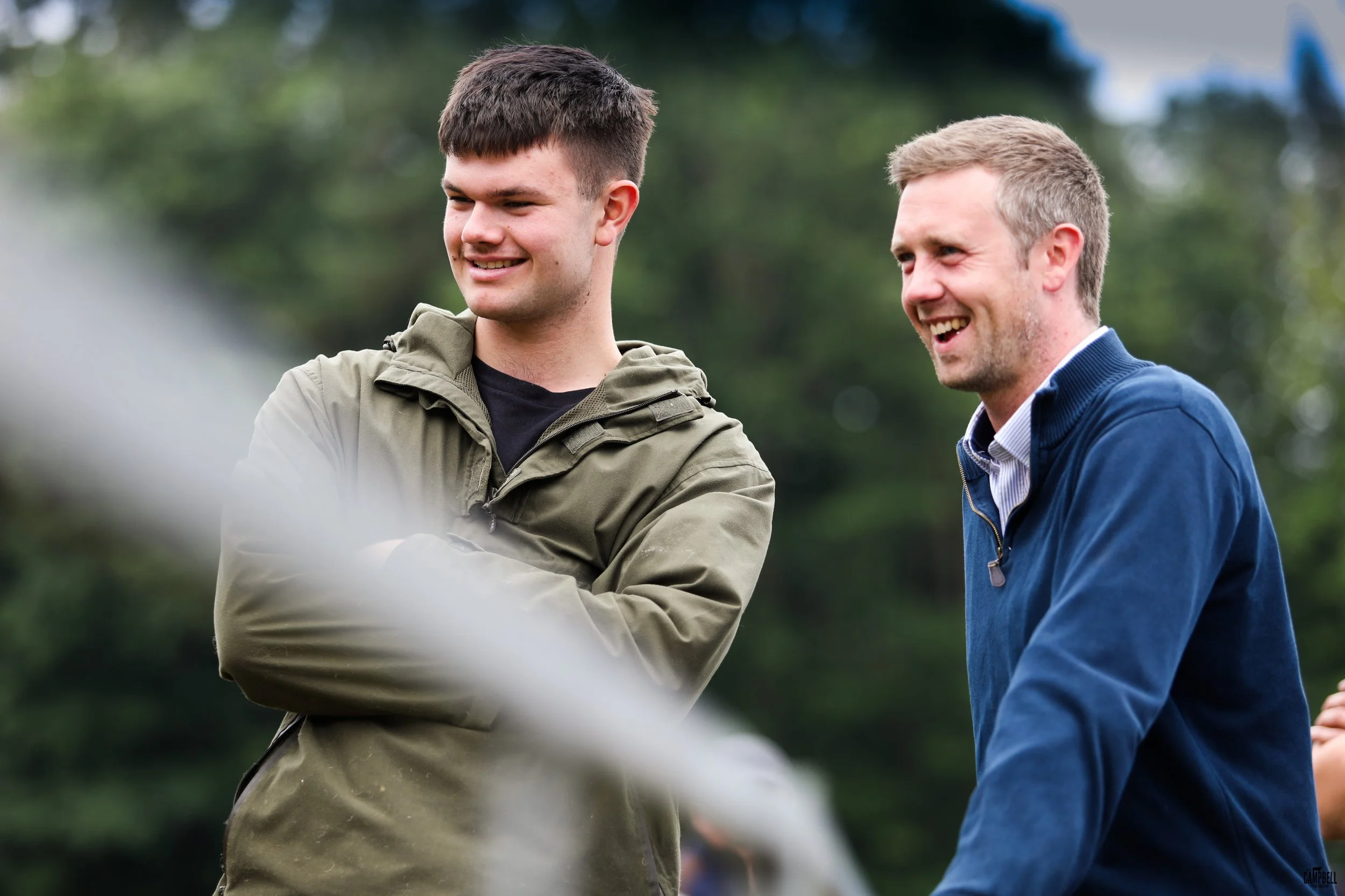 Two young men, one with dark hair and the other with blond hair, smiling and talking outdoors with a blurred green background.