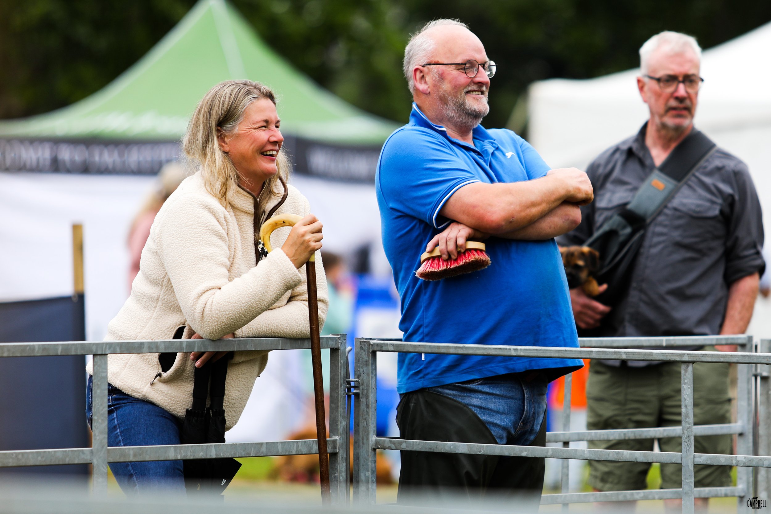 Three people standing behind a metal barrier at an outdoor event, smiling and laughing. A woman on the left is wearing a beige fleece jacket and holding a walking stick, the man in the middle is wearing a blue polo shirt and glasses, with a brush in 