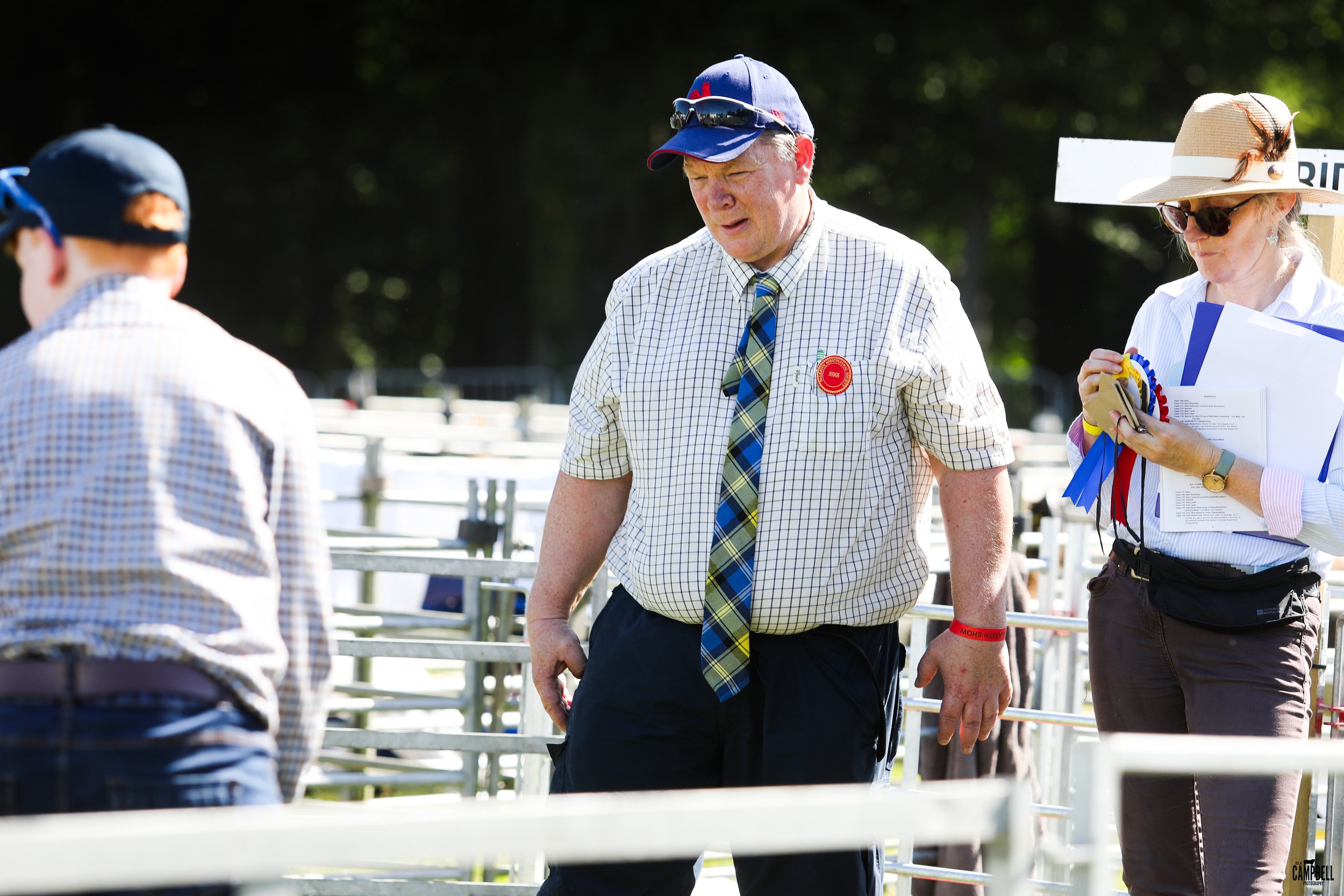 A man in a checkered shirt and tie with a red badge on his chest, standing outdoors among metal fencing, with people and trees in the background.