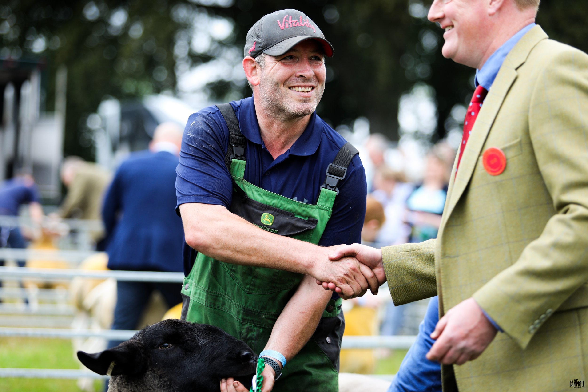 Two men shaking hands at an outdoor event, with a black dog in the foreground and people in the background.