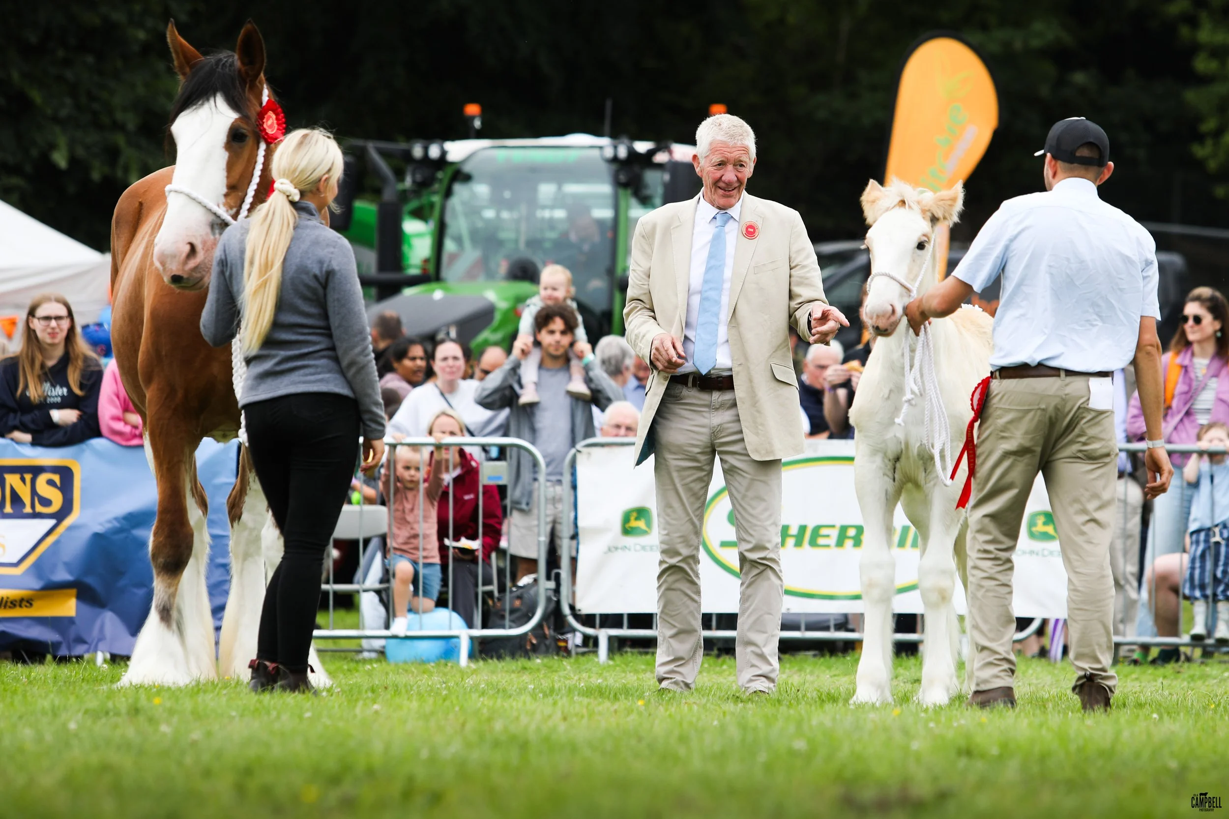 An outdoor horse show with two horses, two handlers, and a judge. The horse on the left is brown and white with a red ribbon on its bridle. The horse on the right is white. The man in the center is smiling and wearing a light-colored suit with a blue