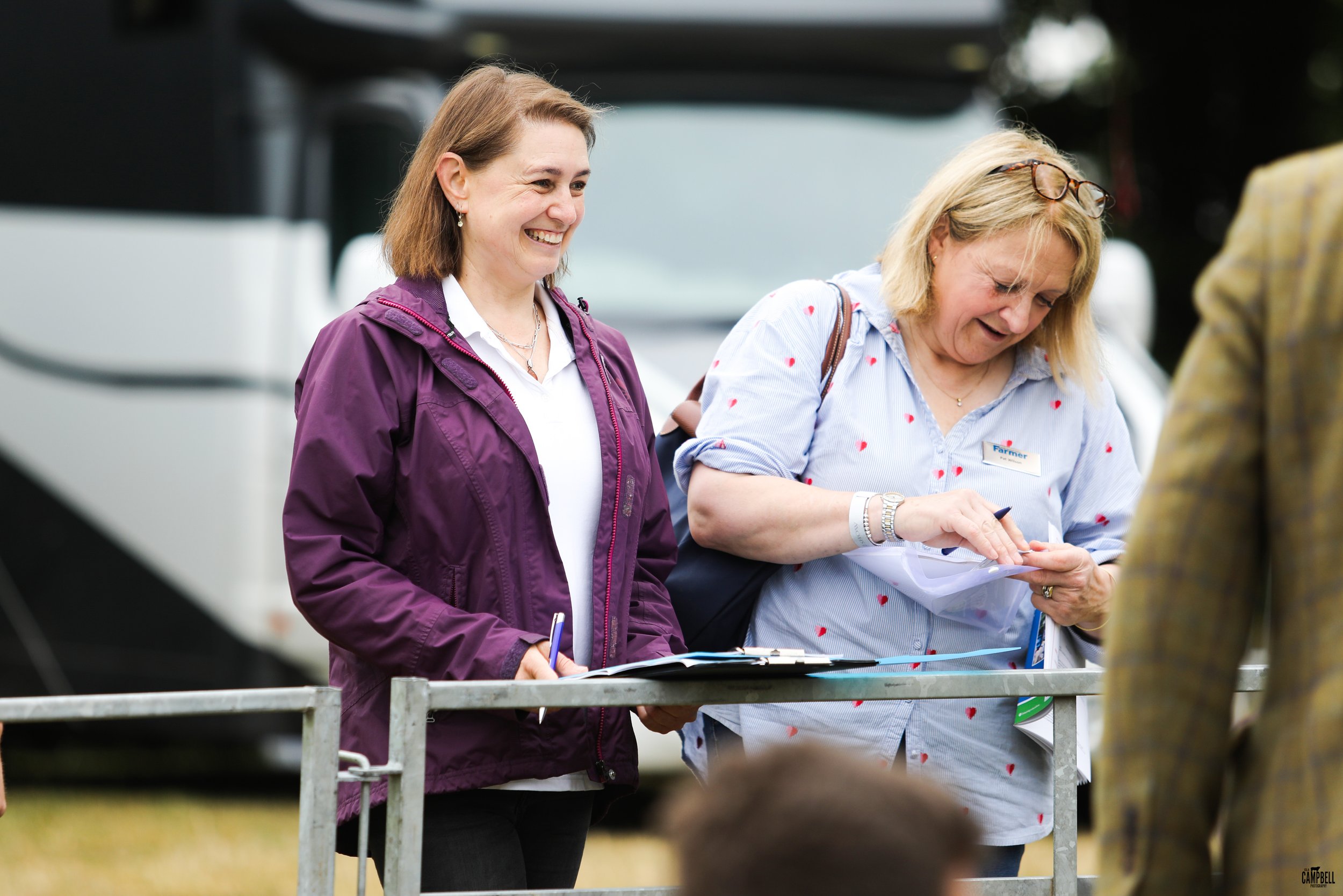 Two women standing outdoors near a railing, one smiling and wearing a purple jacket, the other looking down at papers and wearing glasses on her head, wearing a blue shirt with red hearts.
