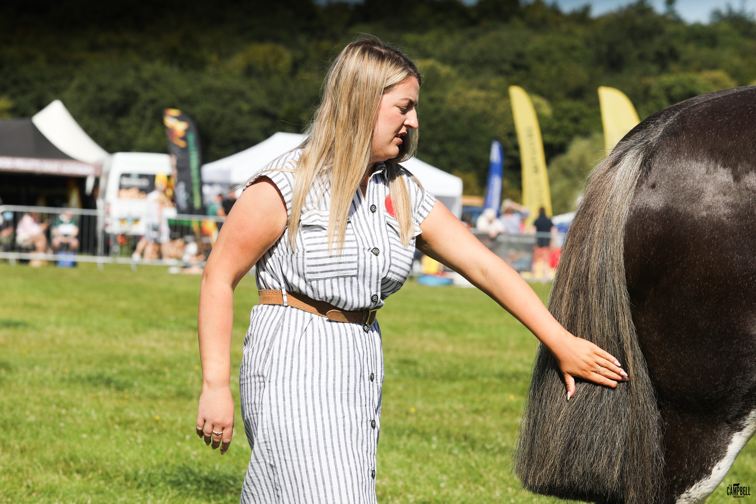 A woman with blonde hair, wearing a striped dress with a brown belt, is petting a black and white horse at an outdoor event on a grassy field.