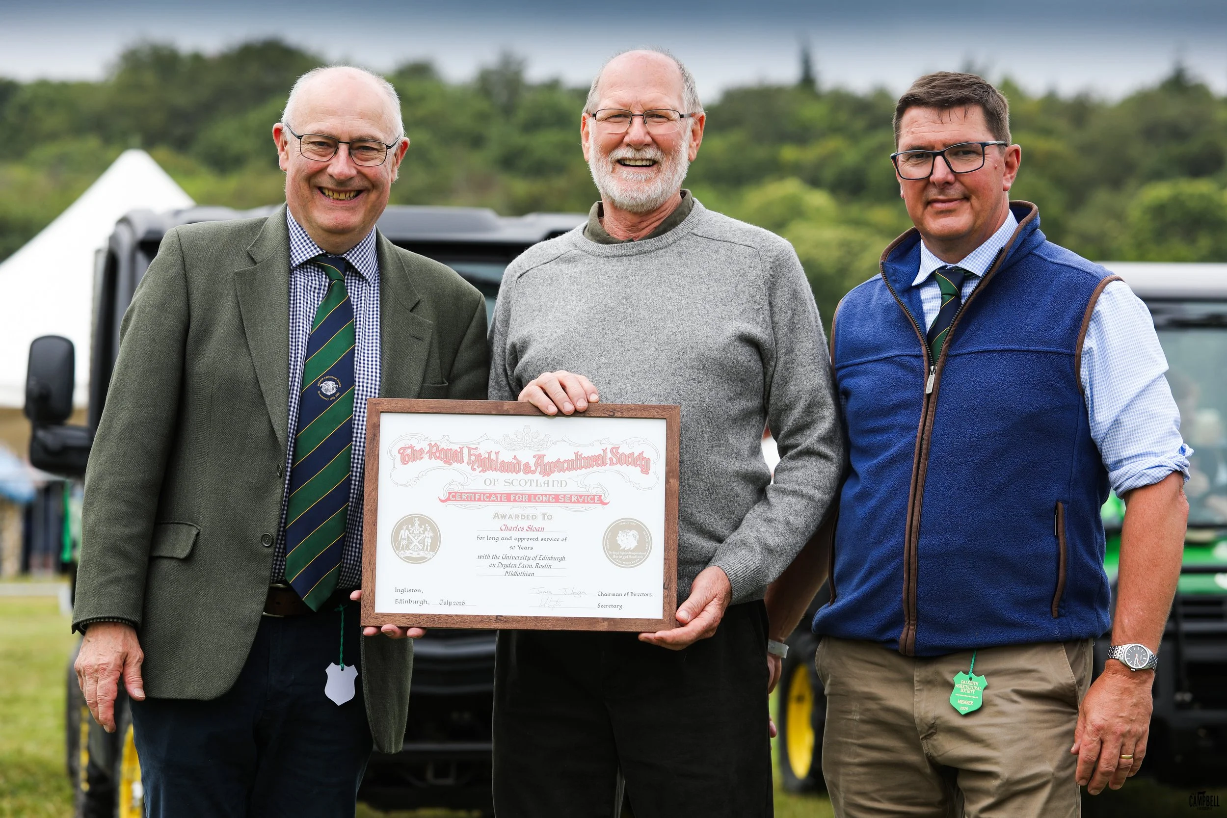 Three men standing outdoors, smiling, one holding a framed certificate for long service award. The man on the left wears glasses, a green blazer, and a tie; the man in the middle has a beard and glasses, and wears a grey sweater; the man on the right