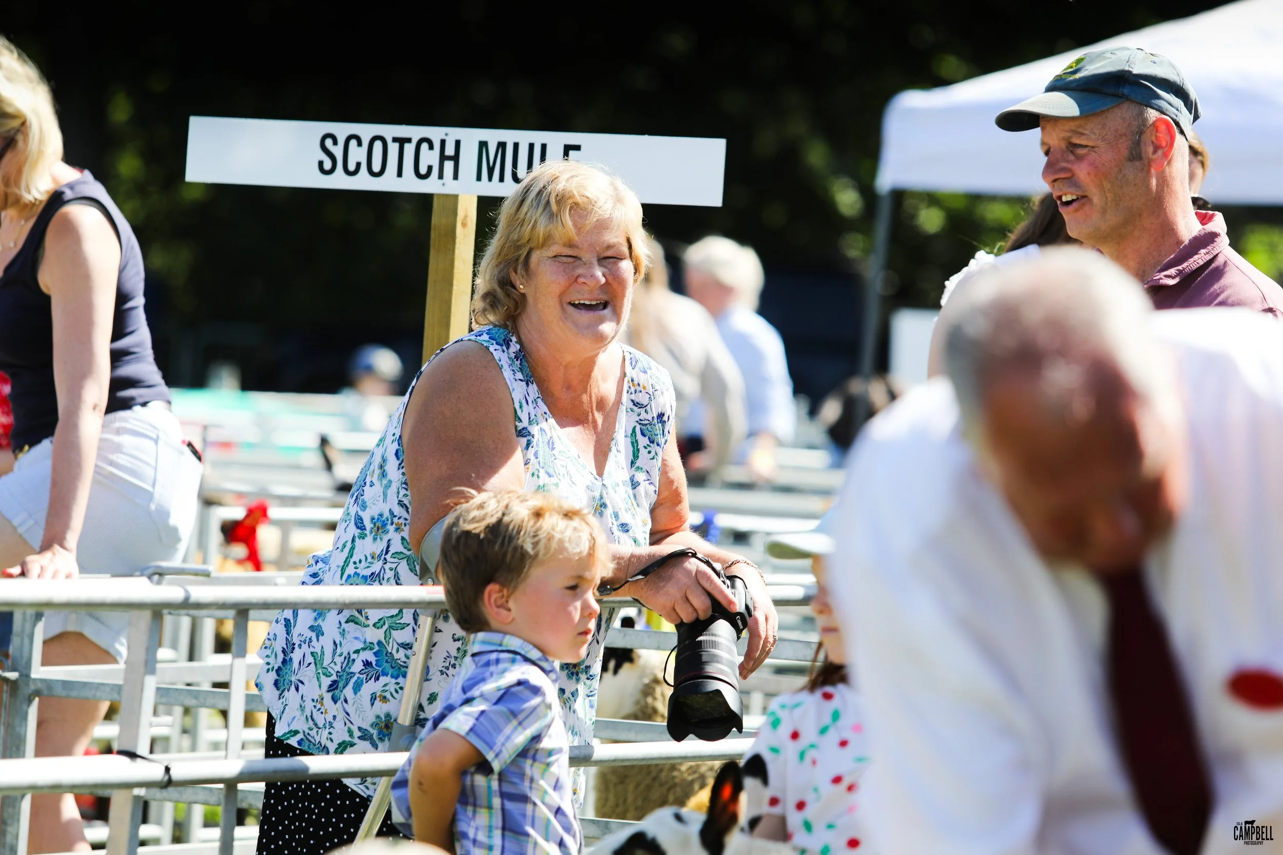 People at a fair or outdoor event, with a woman in a floral top holding a camera and a young boy in plaid standing nearby, and a sign that reads 'Scotch Mule' in the background.
