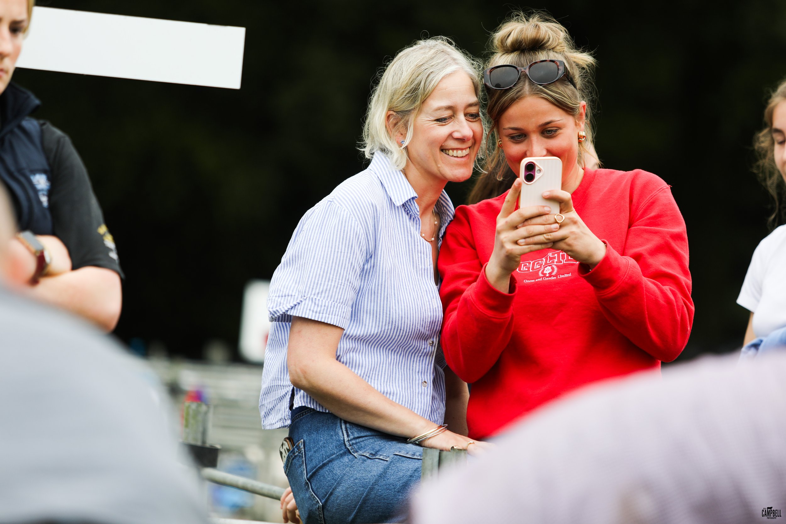 Two women smiling and looking at a smartphone at an outdoor event, with a dark background and several people around.