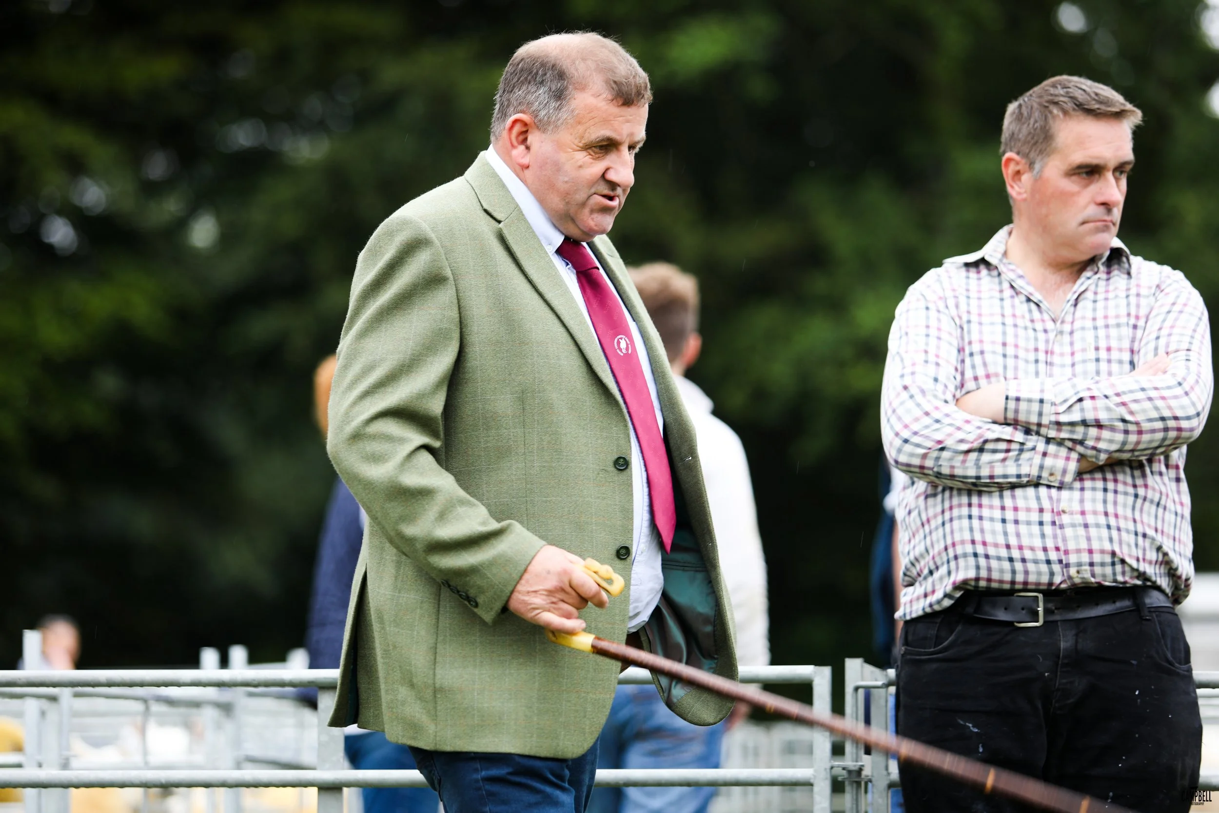 A man with gray hair wearing a green blazer and a burgundy tie standing outdoors, holding a riding crop, with a serious expression, alongside another man in a checkered shirt with arms crossed. The background is green with blurred trees.