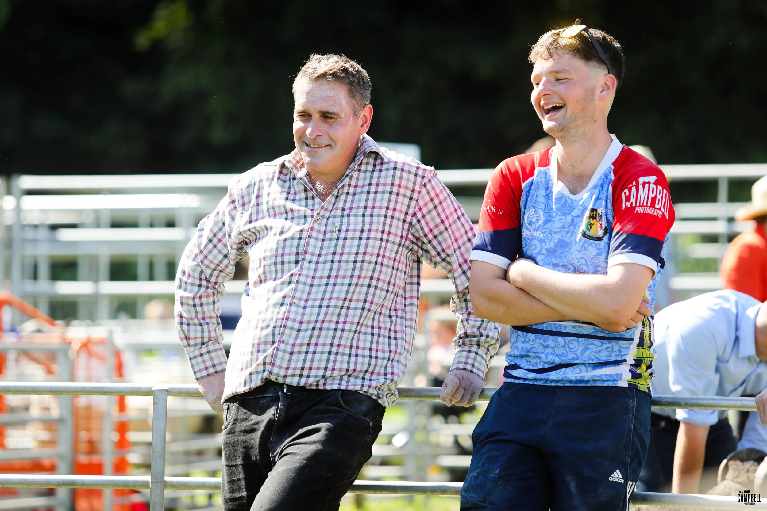 Two men standing outdoors at a sporting event, smiling and talking to each other, with a fence and bleachers in the background.