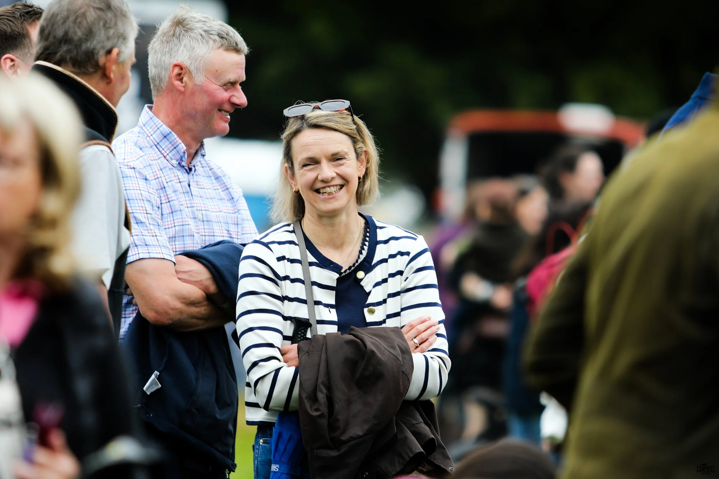 A group of people standing outdoors, smiling and talking. The central woman has blonde hair, sunglasses on her head, and is wearing a navy and white striped sweater. The man next to her has gray hair, a light checked shirt, and is smiling.