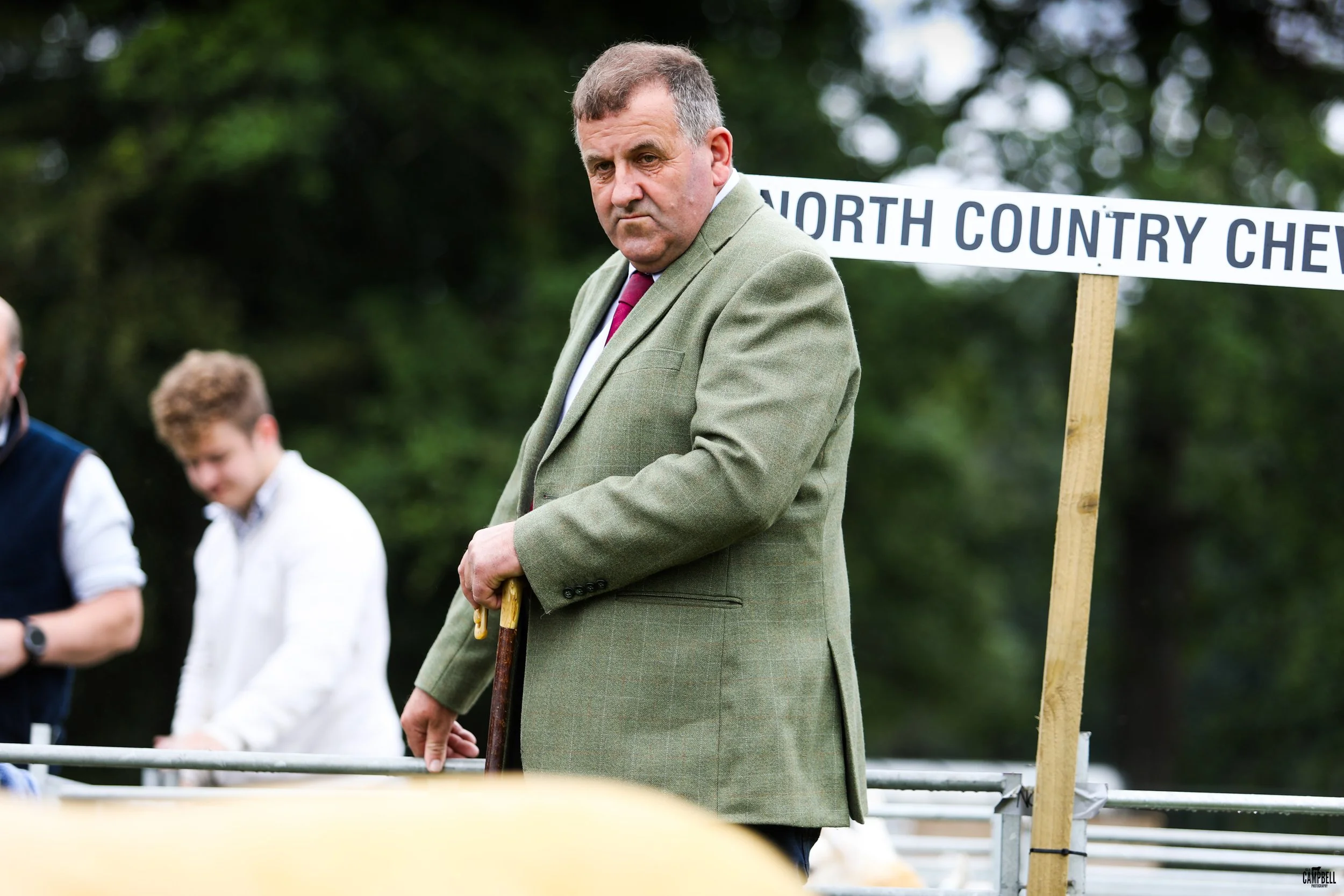 Man in a green blazer holding a cane, standing outdoors near a sign that reads 'NORTH COUNTRY CHE' with trees in the background.