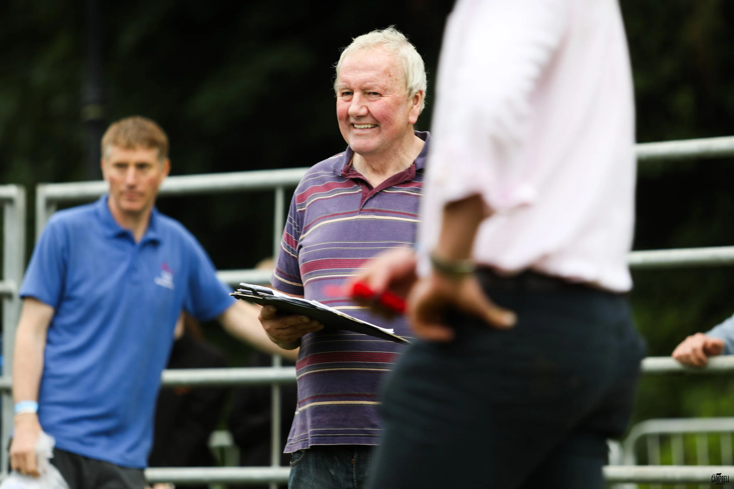 A smiling elderly man with white hair wearing a striped polo shirt holding a clipboard, engaging in conversation with a person in a white shirt, while other people listen in the background.