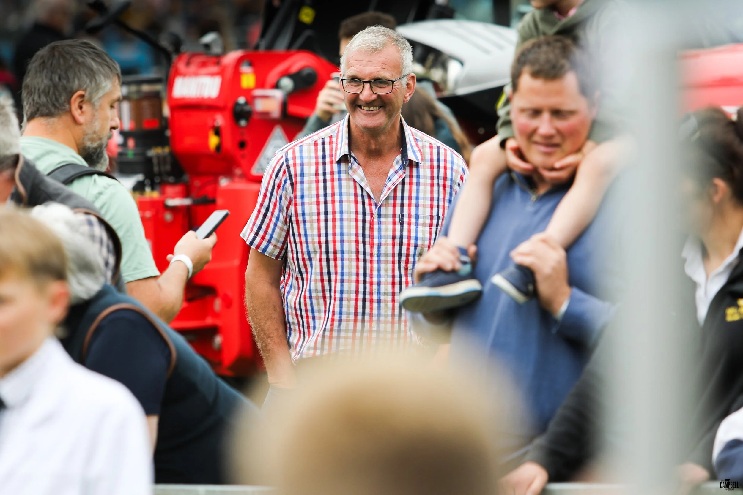 A group of people gathered at an outdoor event, with a smiling man in glasses and a checkered shirt engaging with others. The background shows red machinery and various attendees.