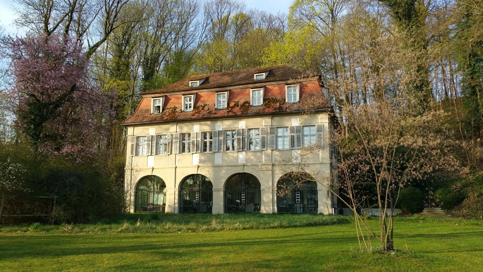 Frontfassade eines historischen Gebäudes, das ebenerdig vier große Rundbögen besitzt, die mit Glas geschlossen wurden. Darüber ein Geschoss mit je zwei Fenstern über einem Rundbogen. Darüber ein Mansarddach mit 5 Gauben und 2 Gauben im Spitzboden.