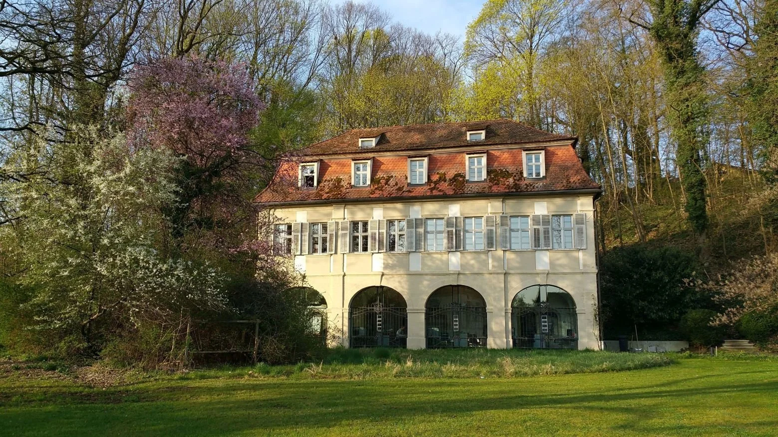 Frontfassade eines historischen Gebäudes, das ebenerdig vier große Rundbögen besitzt, die mit Glas geschlossen wurden. Darüber ein Geschoss mit je zwei Fenstern über einem Rundbogen. Darüber ein Mansarddach mit 5 Gauben und 2 Gauben im Spitzboden.