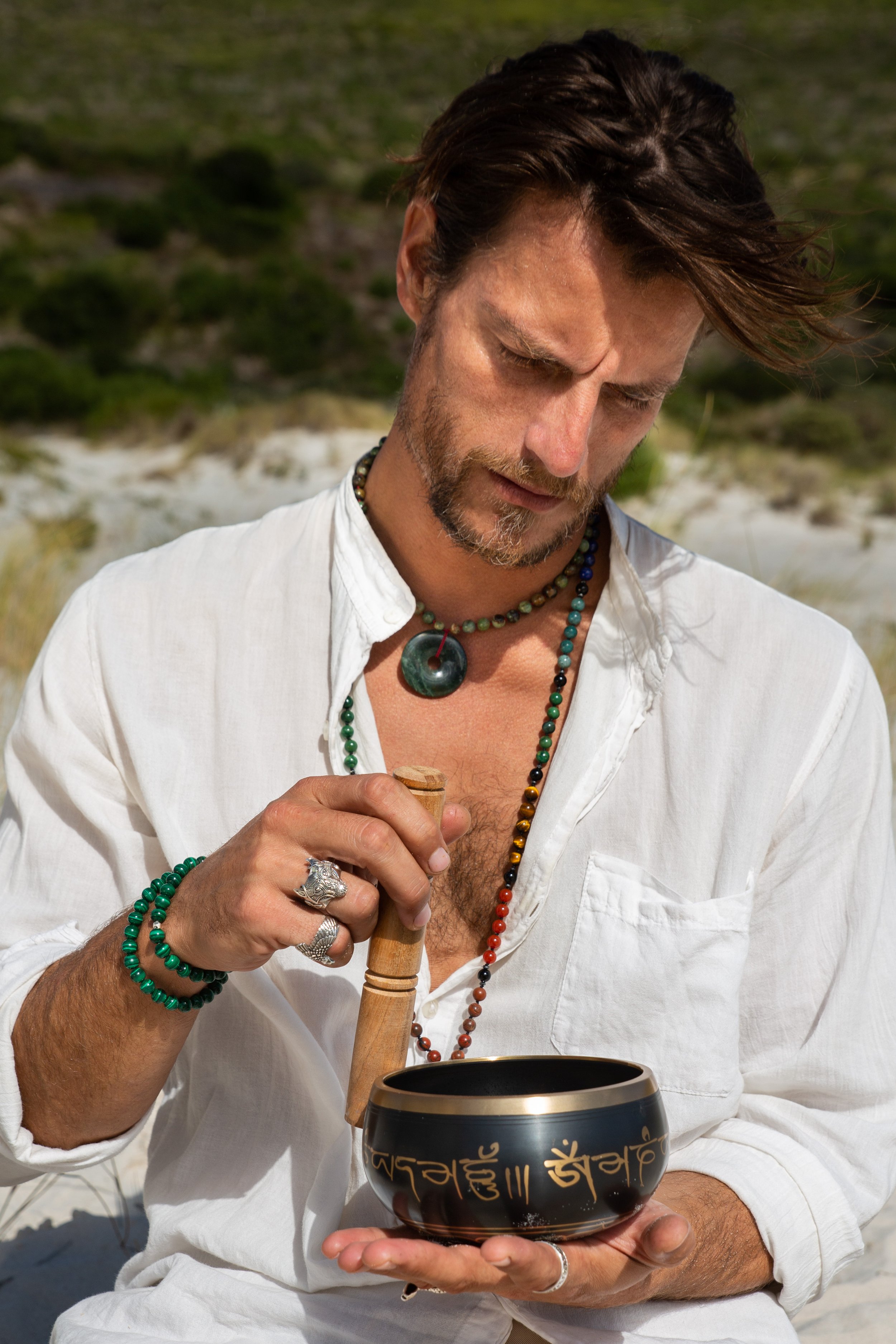 A man in a white shirt playing a Tibetan singing bowl outdoors, wearing colorful bead necklaces and rings, with a natural background of sand and greenery.