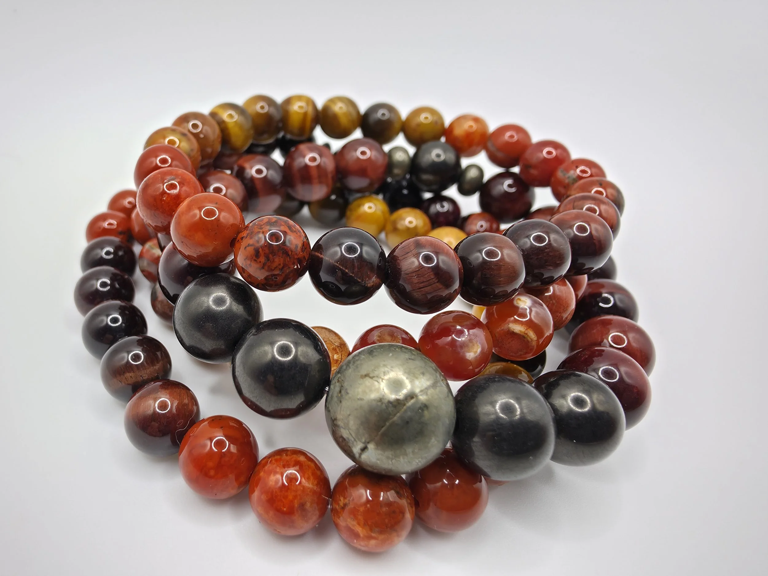 Three beaded bracelets made of polished, multicolored round stones, including red, black, brown, and green, arranged on a white surface.