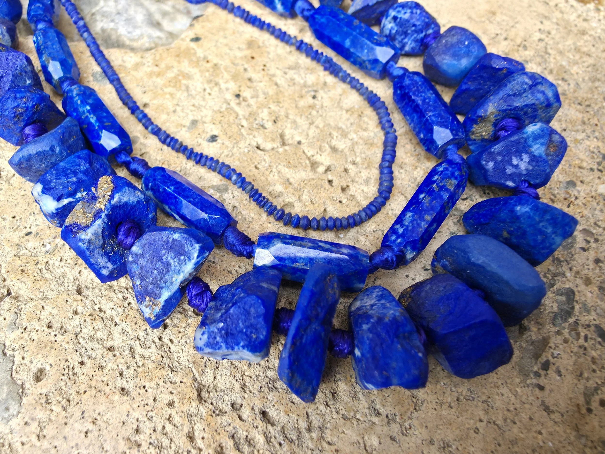 Close-up of multiple layered blue beaded necklaces with irregular blue stone beads on a sandy surface.