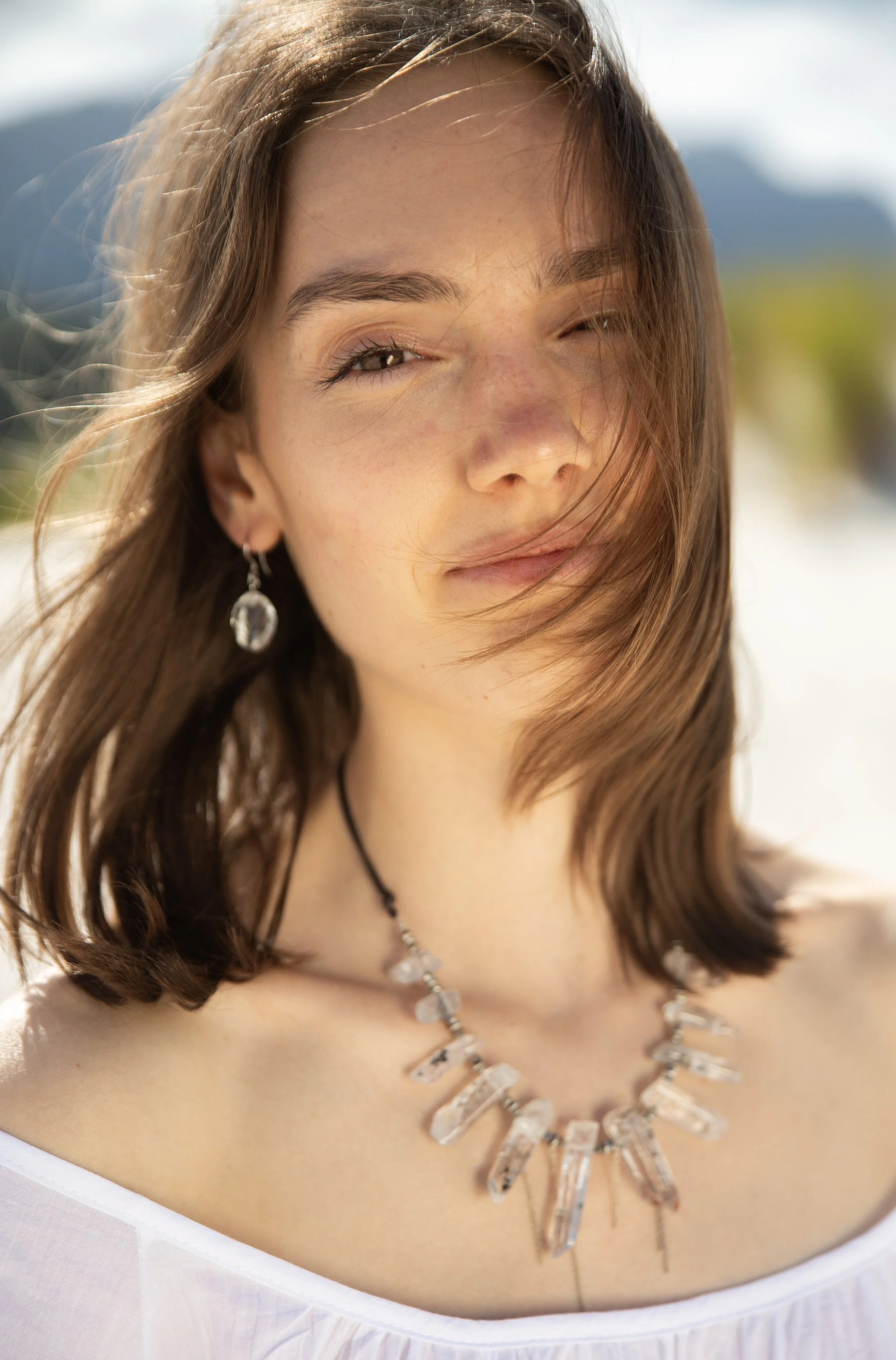 A young woman with brown hair, one eye closed, smiling slightly, outdoors in sunlight, with a blurred background of nature.