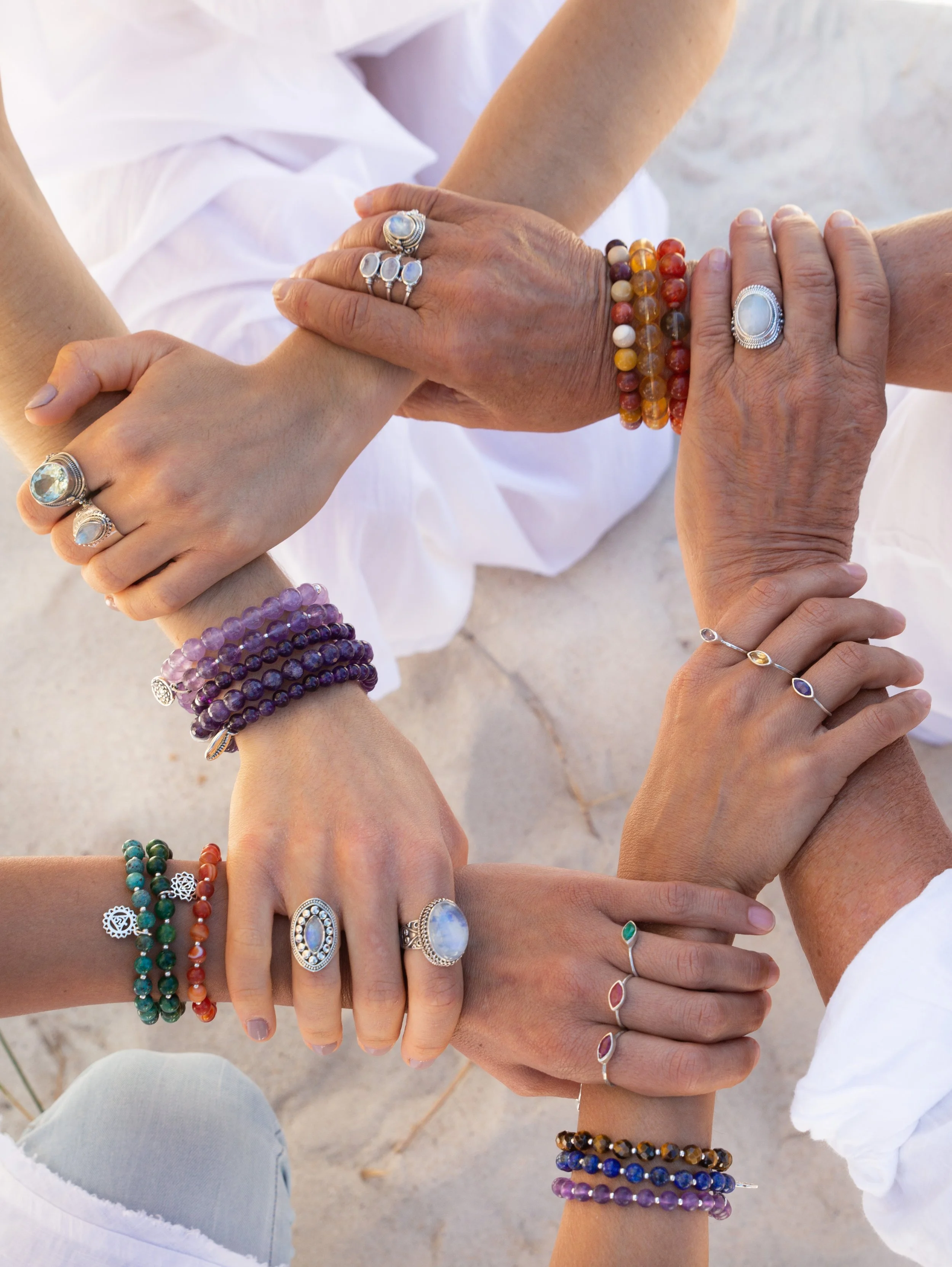 Multiple hands with colorful gemstone and beaded jewelry clasping in a circle on a sandy surface.