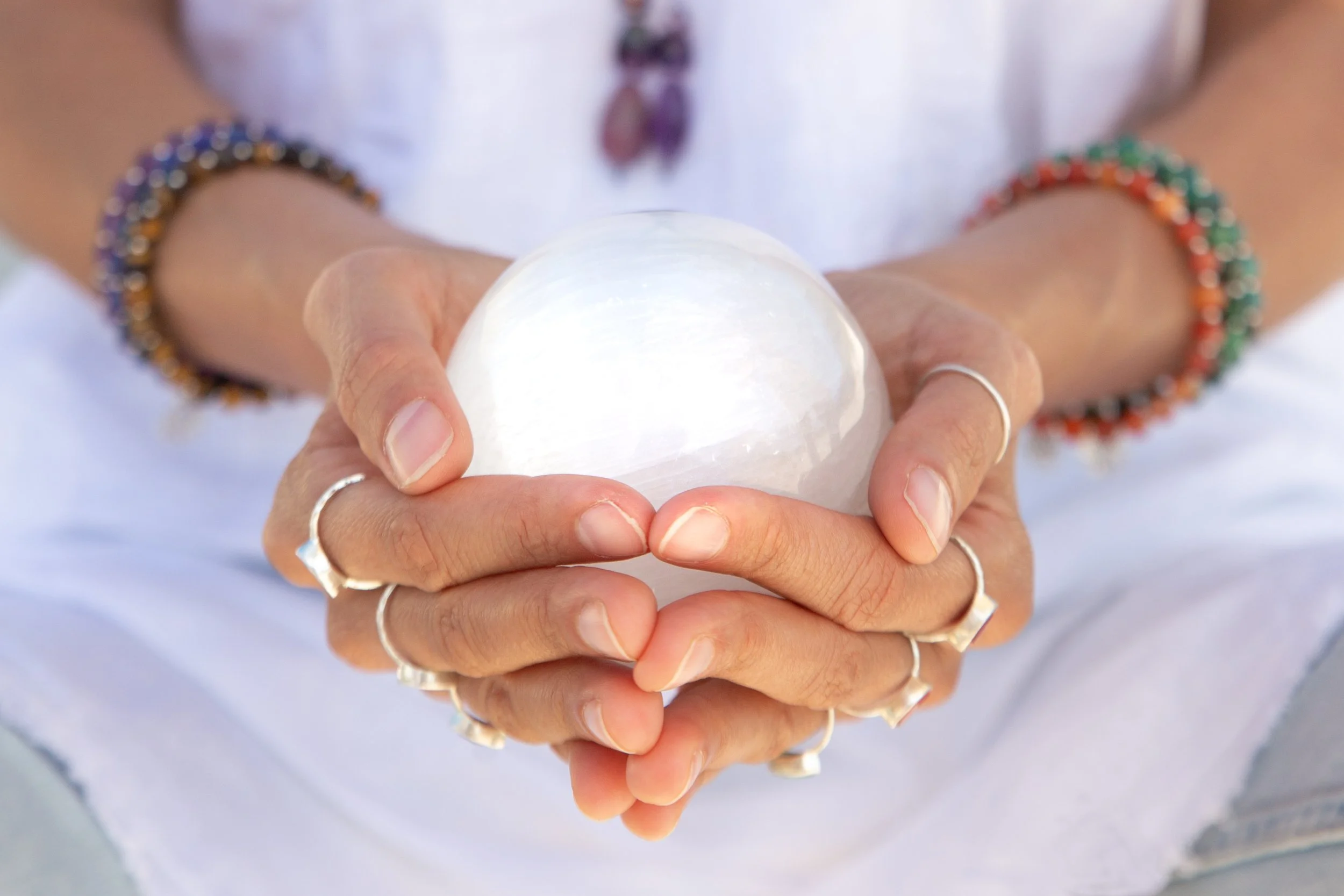 Person holding a glowing white crystal ball with hands adorned with rings and beaded bracelets.