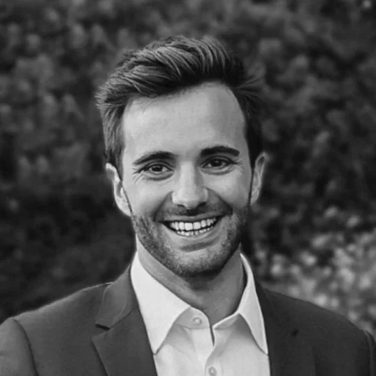 Un homme souriant en costume, photo en noir et blanc, arrière-plan flou.