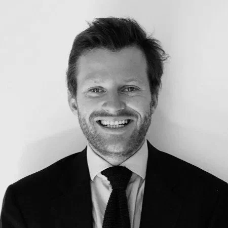 Un homme souriant avec des cheveux courts et une barbe, portant un costume noir et une cravate, contre un fond blanc.