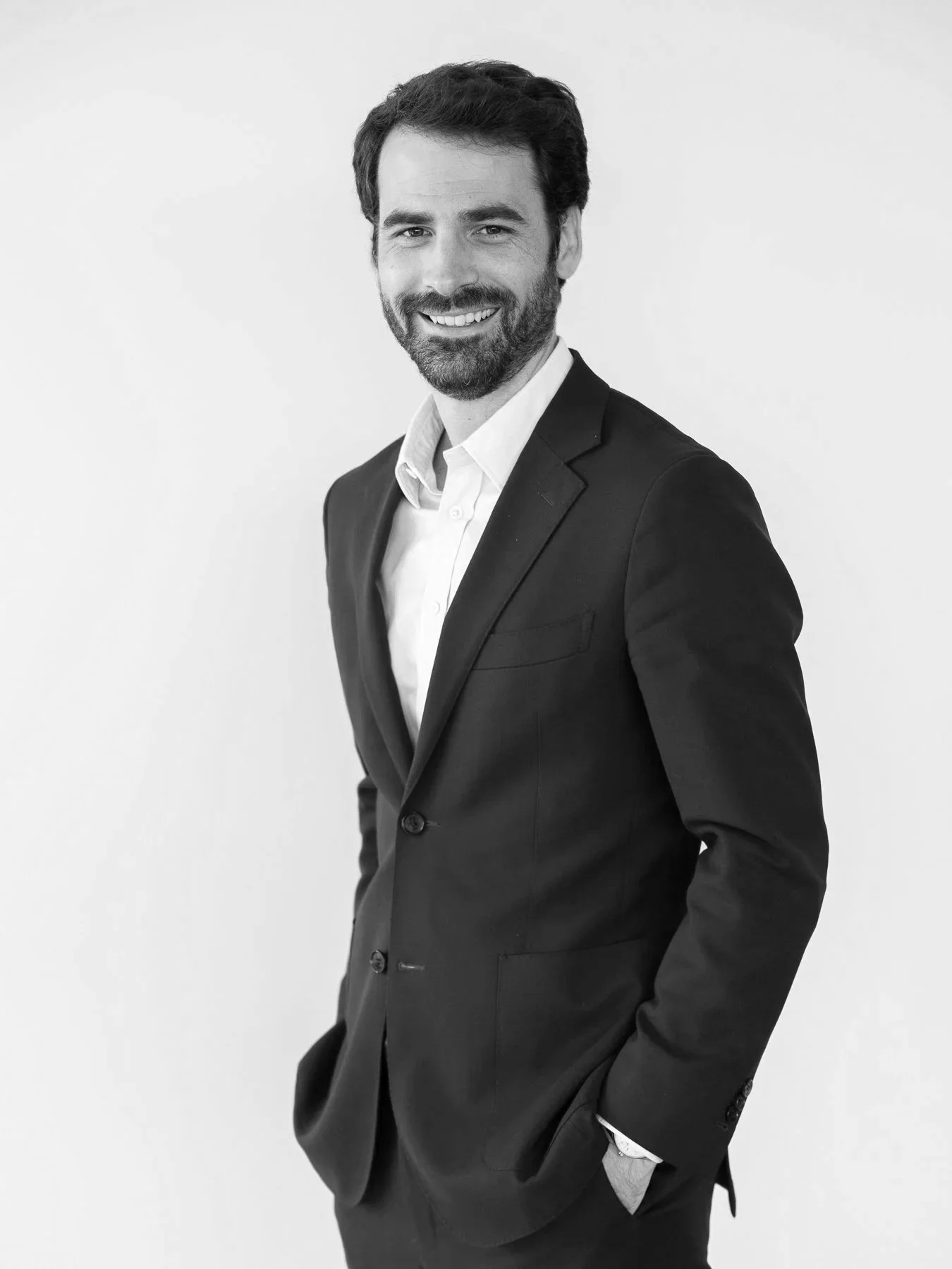 Un homme souriant en costume noir et chemise blanche, posant contre un fond blanc.