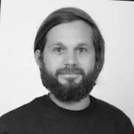 Photo en noir et blanc d'un homme avec une barbe et des cheveux courts, portant un t-shirt noir, souriant face à la caméra.