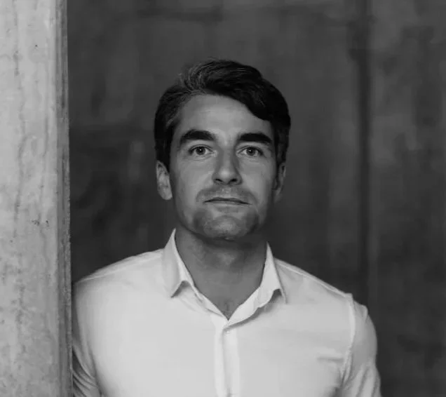 Un homme en chemise blanche qui regarde vers la caméra, debout près d'un mur en pierre, avec un arrière-plan flou.