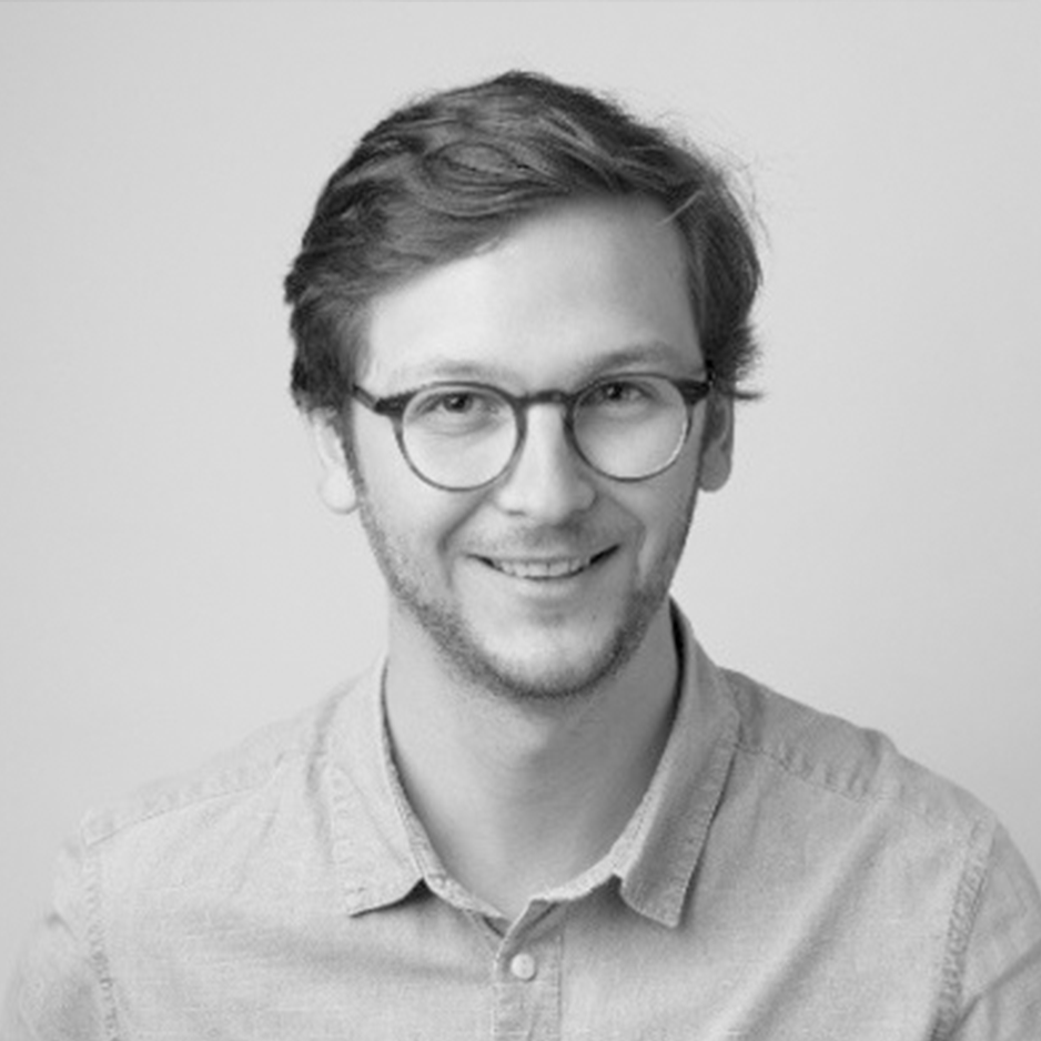 Portrait en noir et blanc d'un jeune homme portant des lunettes, souriant, avec une chemise à col. Fond blanc unicolore.