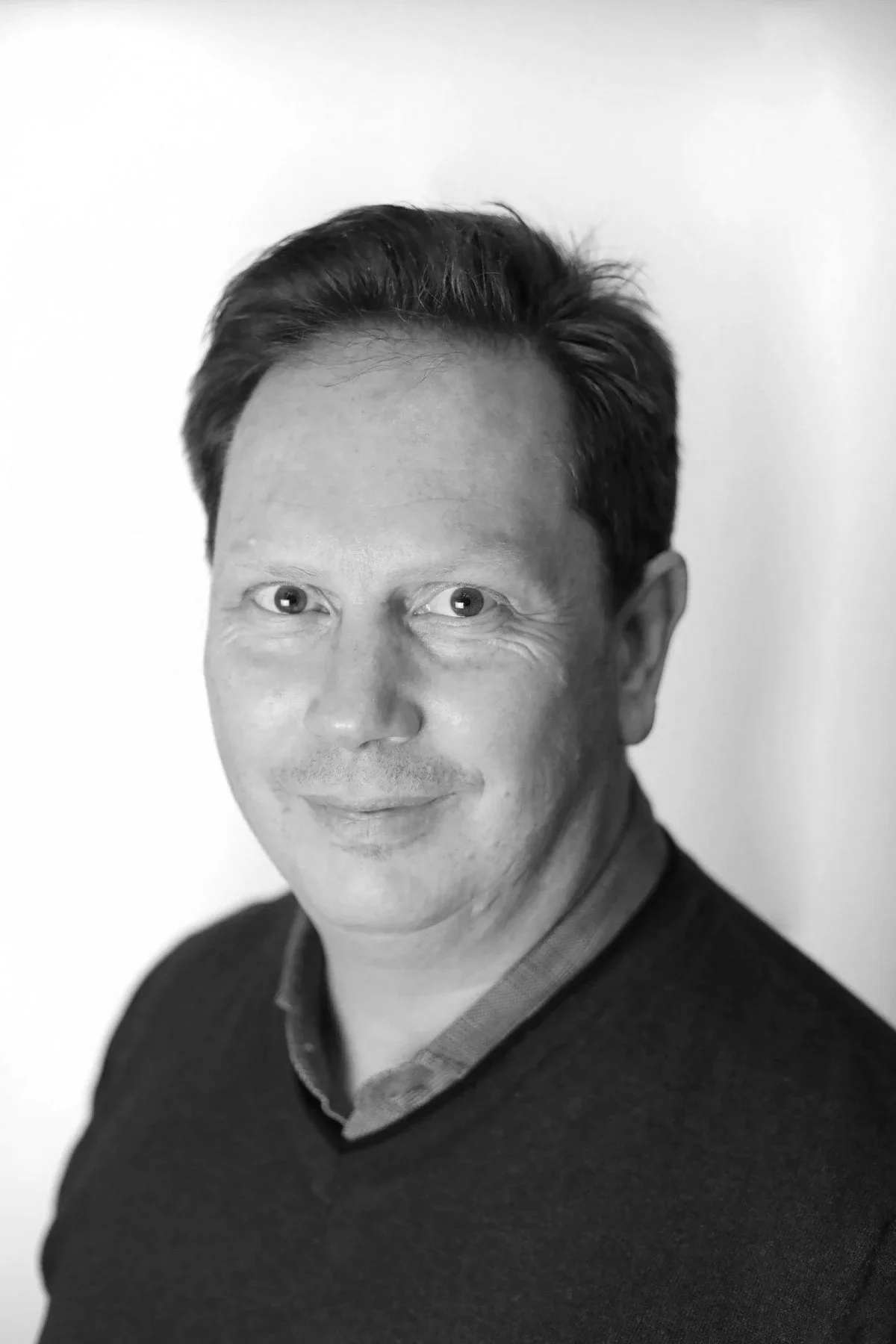 Portrait en noir et blanc d'un homme souriant, portant un pull foncé sur une chemise à col. Fond neutre.